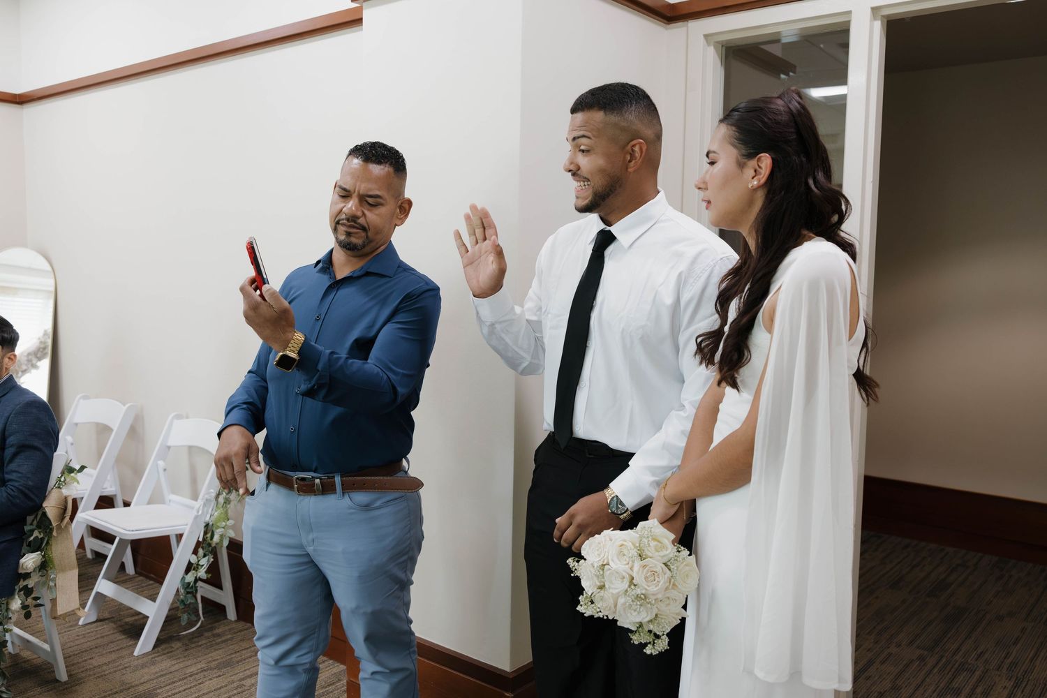 A groom in white shirt and black tie raises his hand beside a bride holding white flowers as a man photographs them.