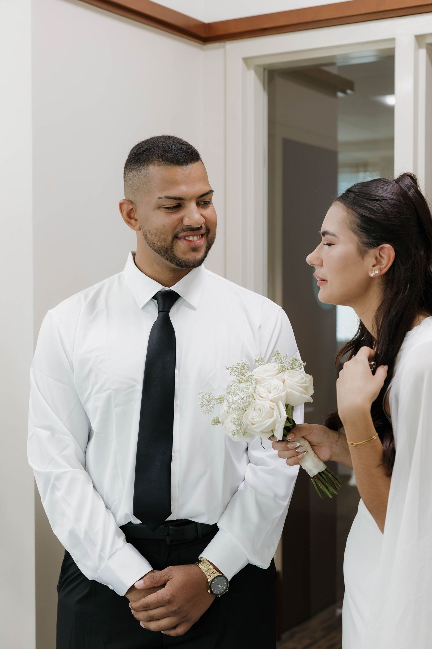 Groom in white shirt and black tie smiles at bride holding white rose bouquet during wedding day preparations.