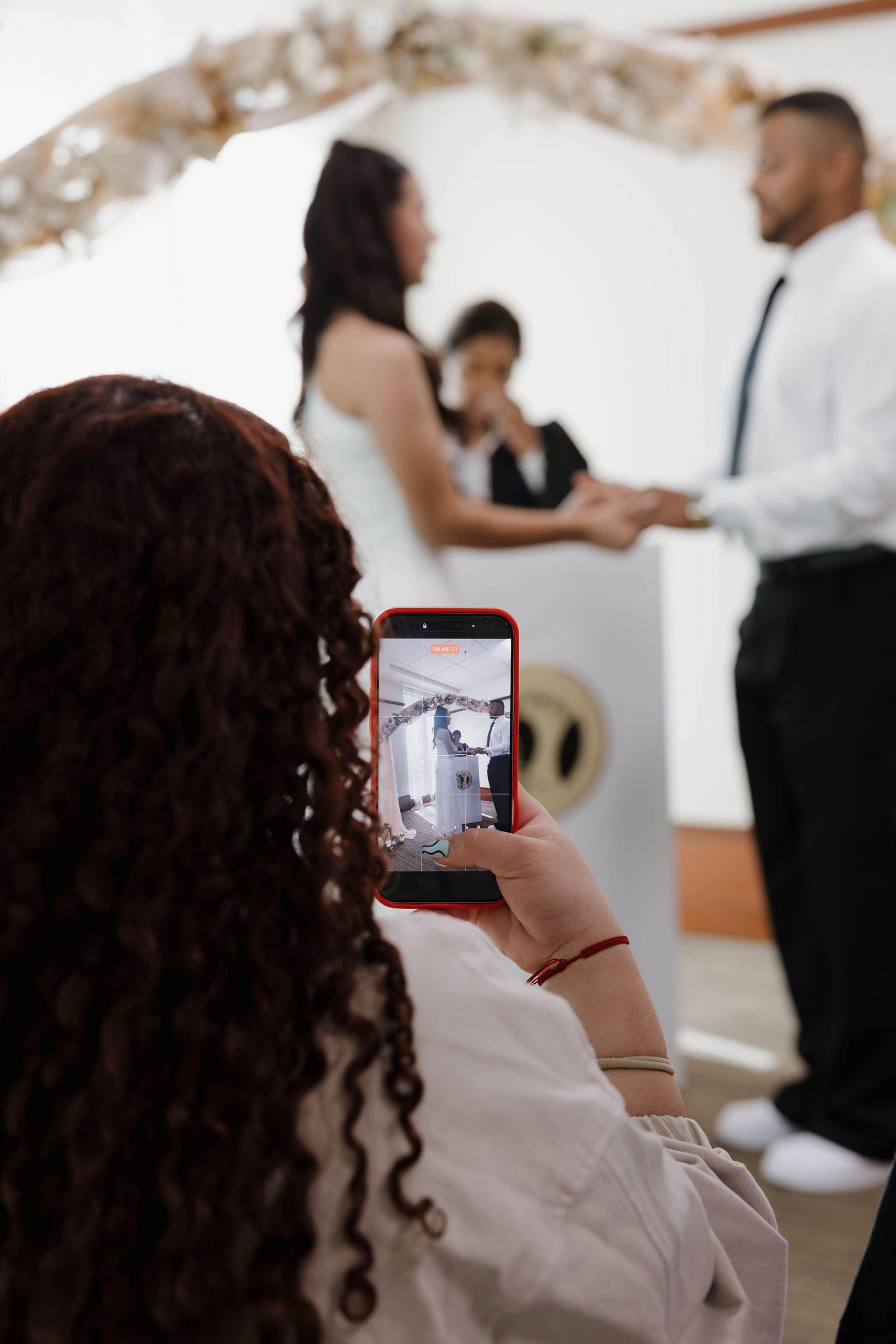 Woman with curly red hair photographs a wedding ceremony on her smartphone with a red case.
