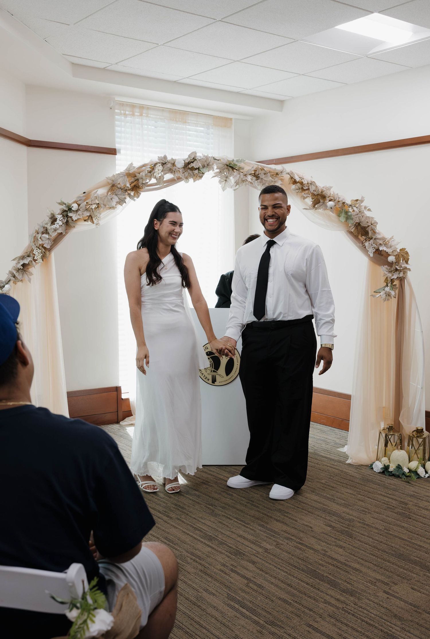 Newlywed couple holds hands smiling under floral arch after wedding ceremony in simple indoor venue.