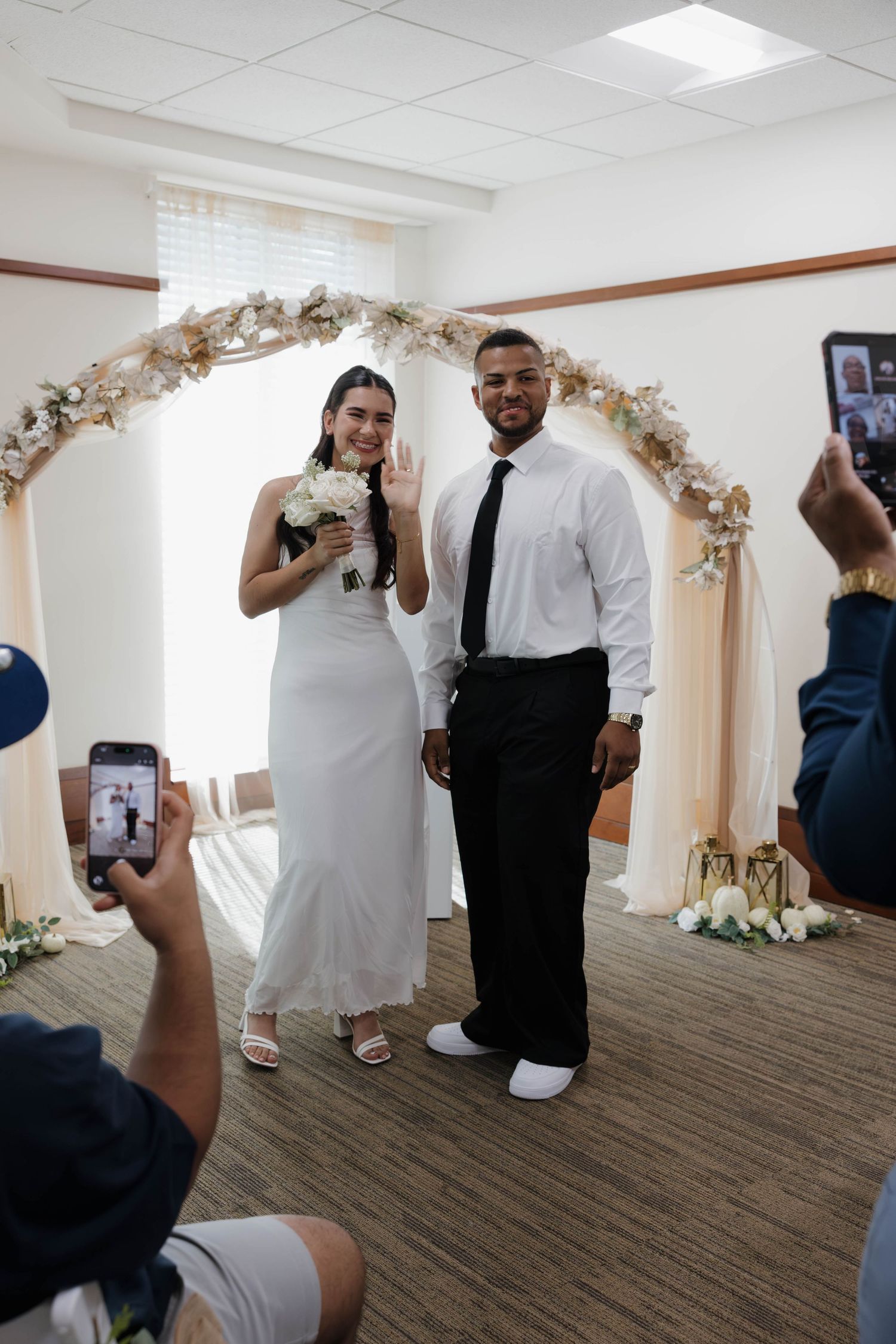 Newlywed couple smiling under floral arch at indoor wedding ceremony while guests photograph them.