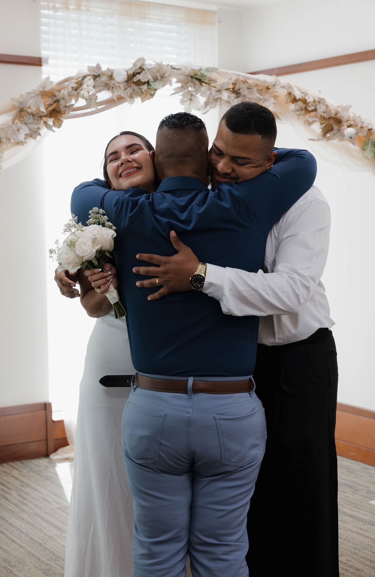 Bride and groom share joyful group hug with guest under floral arch at intimate wedding ceremony.