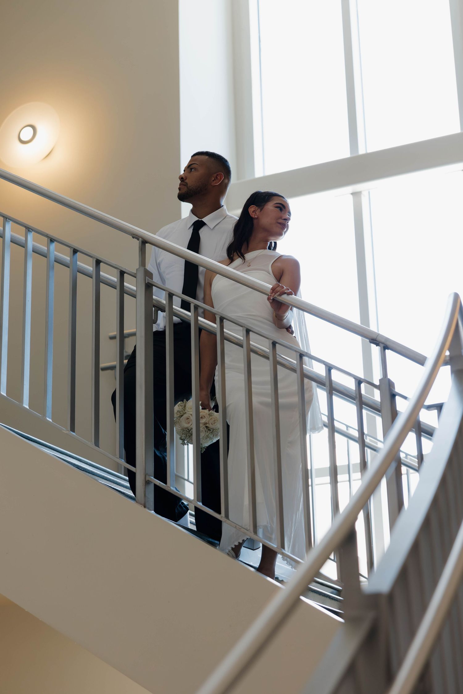 Bride and groom standing together on elegant staircase, woman in white dress holding bouquet, man in black tie attire.