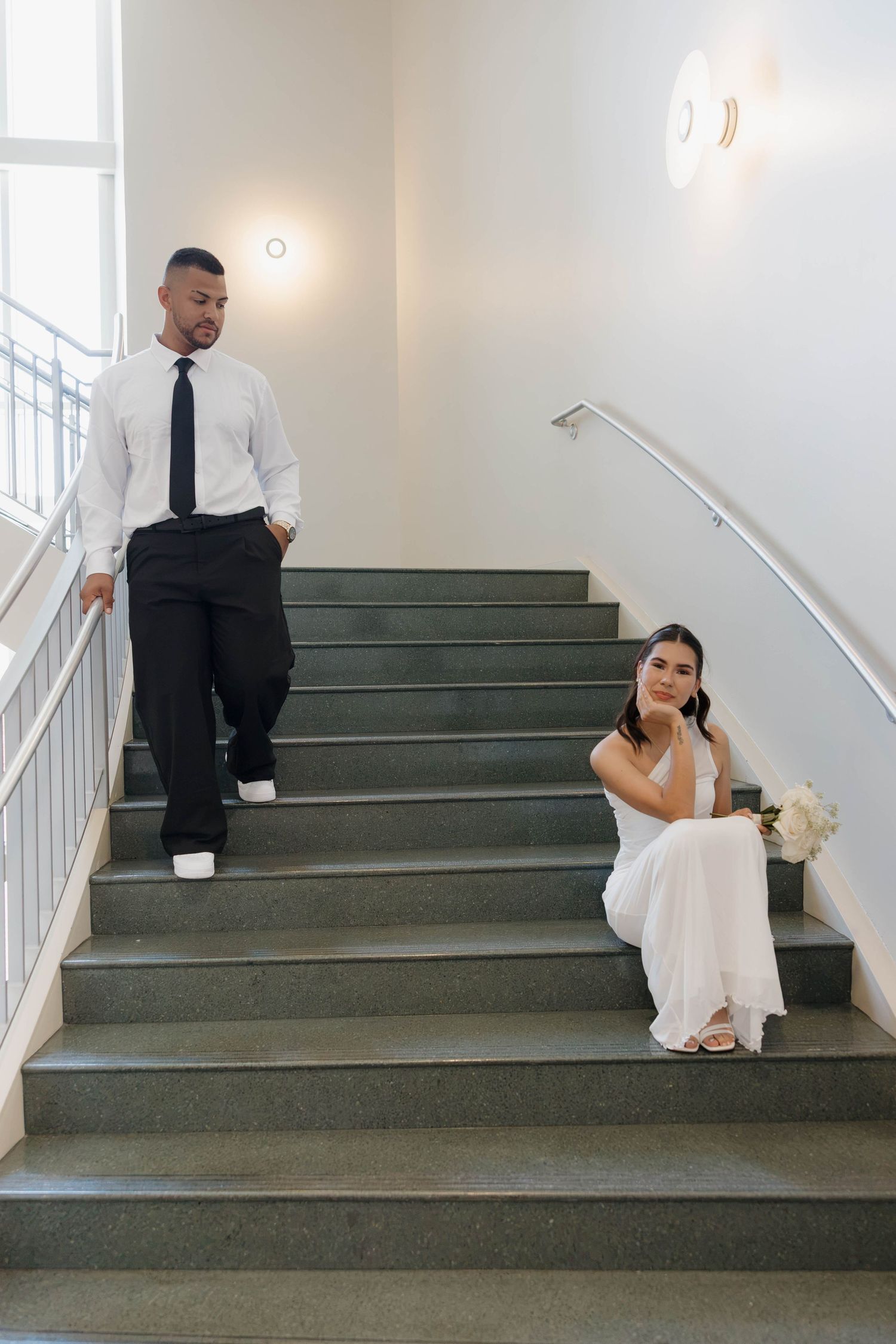 A bride in a white dress sits on stairs while a groom in a white shirt and tie walks down toward her.