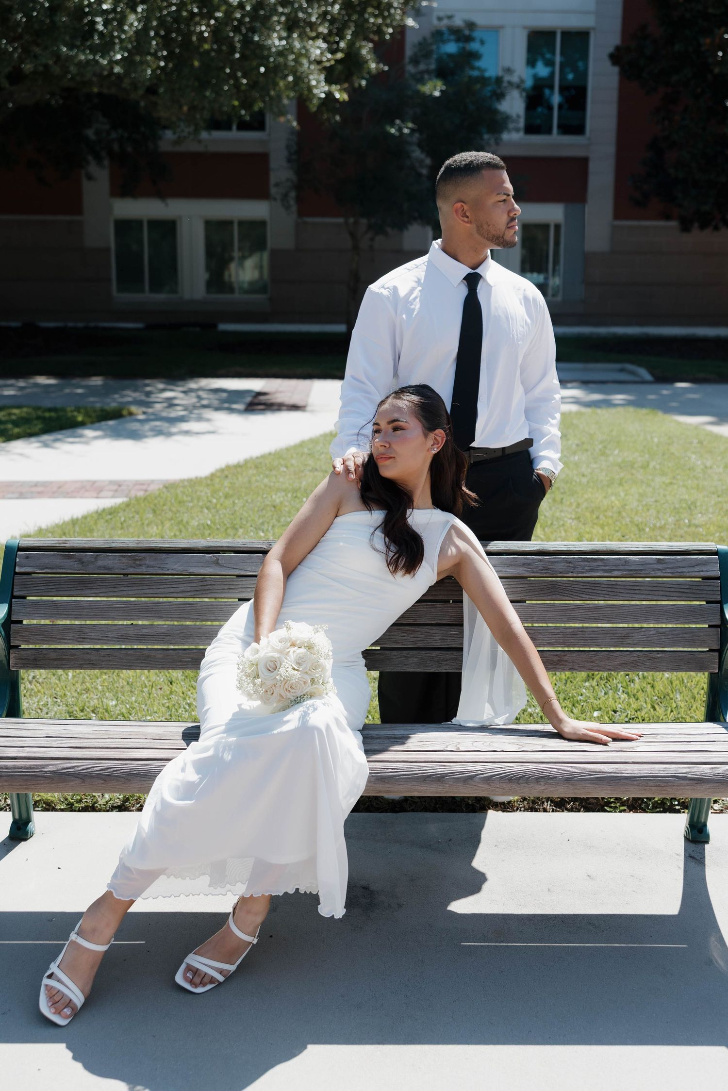 Bride in white dress sits on bench holding bouquet while groom in white shirt and black tie stands behind her outdoors.