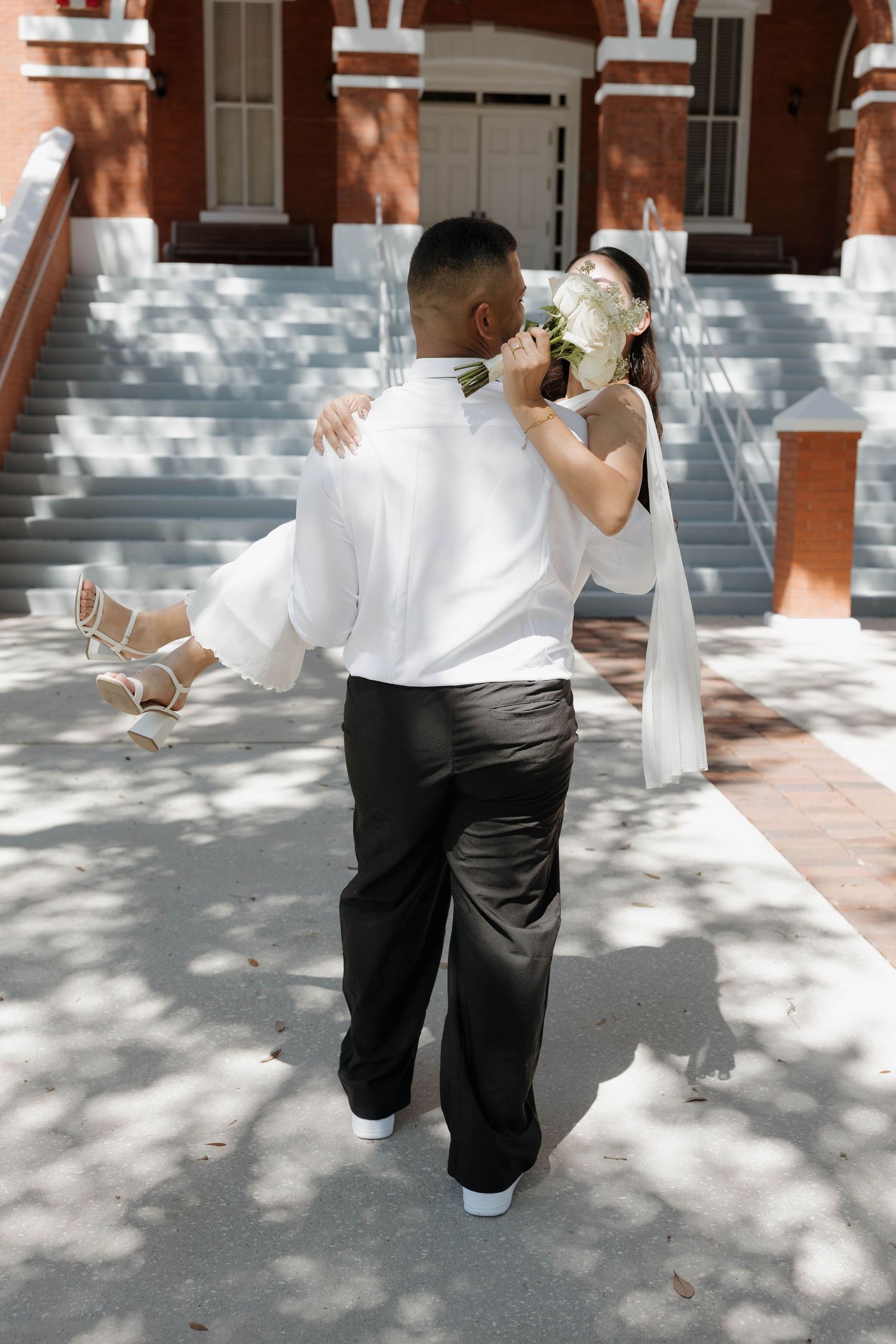 Groom lifts bride outside a brick church as she holds her white bouquet on their wedding day.