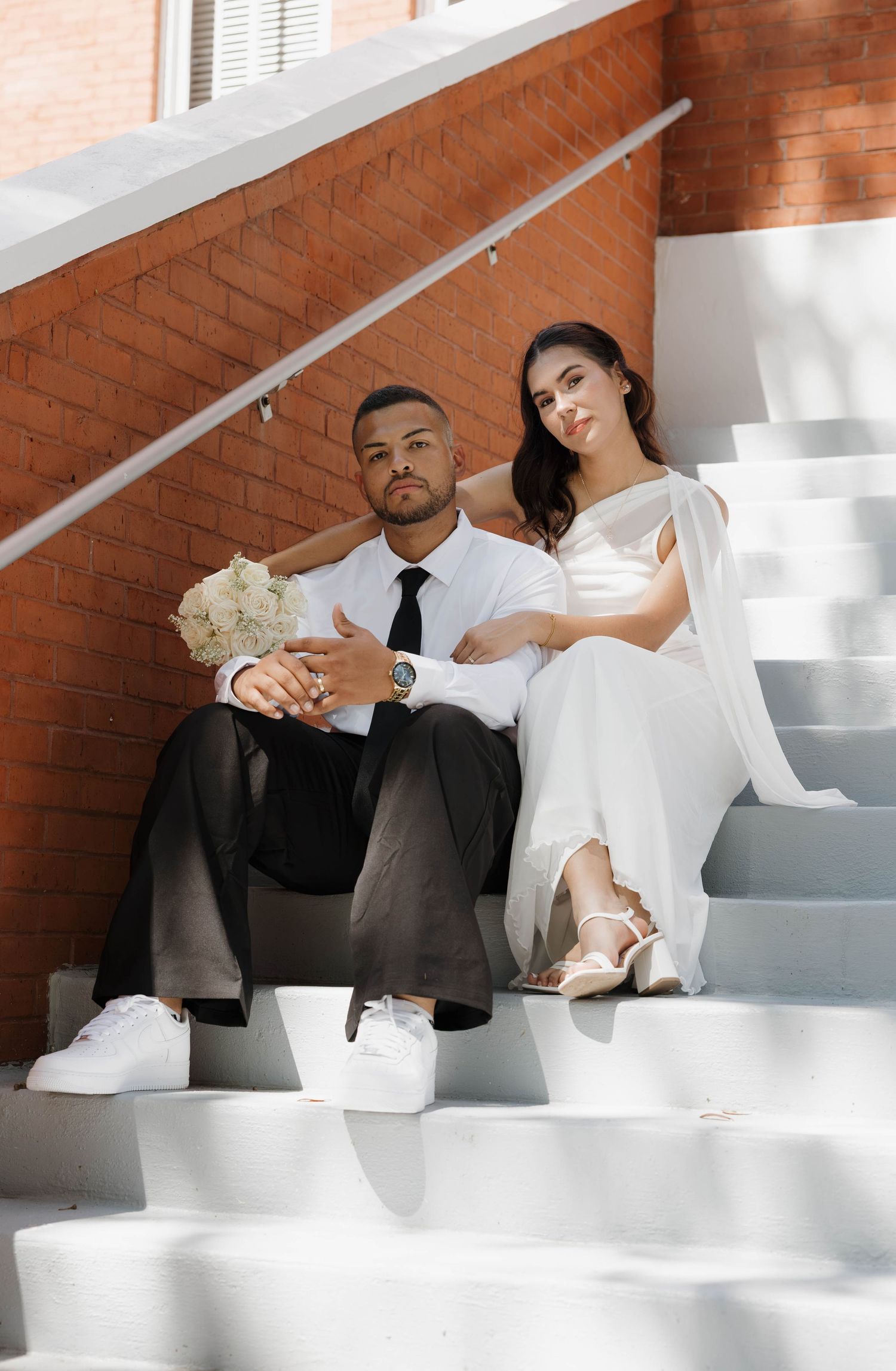 Wedding couple posing on white outdoor steps with bride holding bouquet against brick wall.