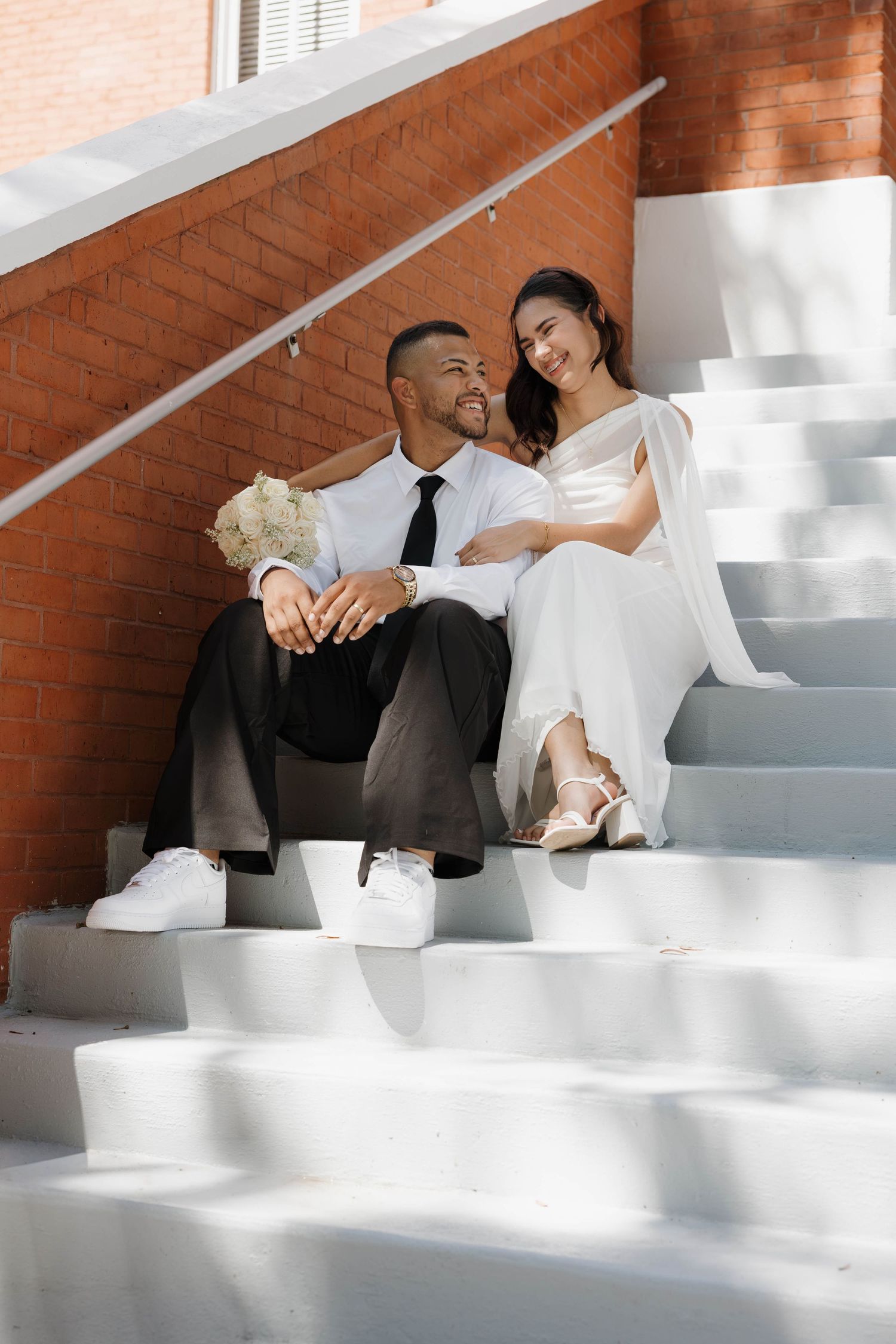 Smiling newlyweds sit together on white outdoor steps holding a white bridal bouquet.
