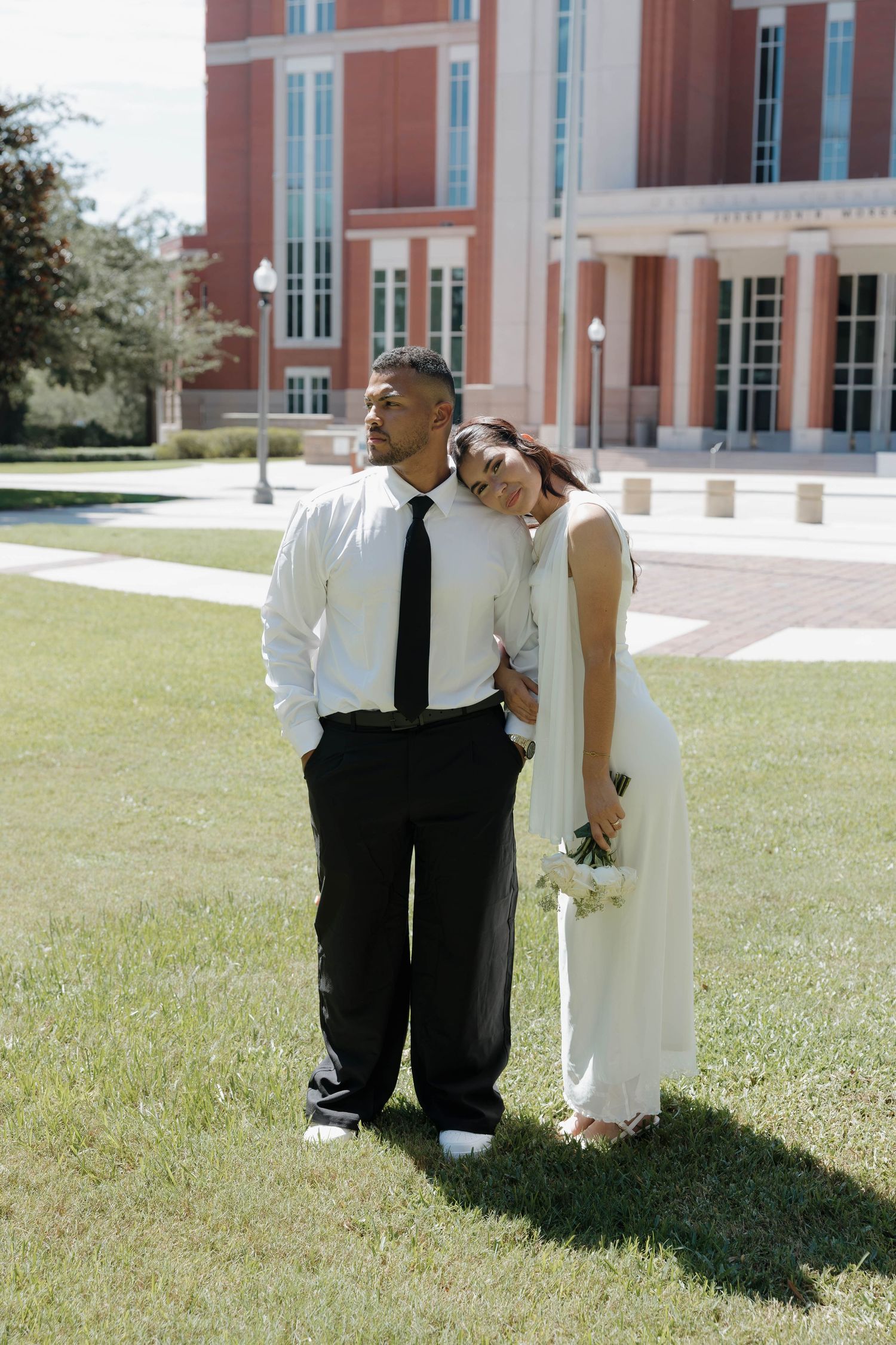 Bride and groom pose together on green lawn outside red brick building, she leans on his shoulder in white dress.