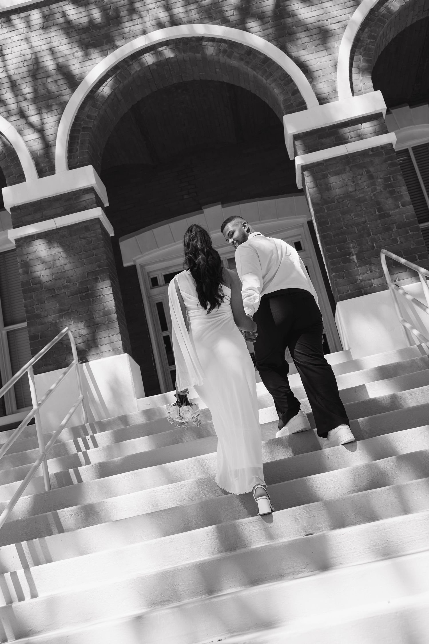 Bride and groom walk hand in hand up steps toward a brick building in a black and white photo.