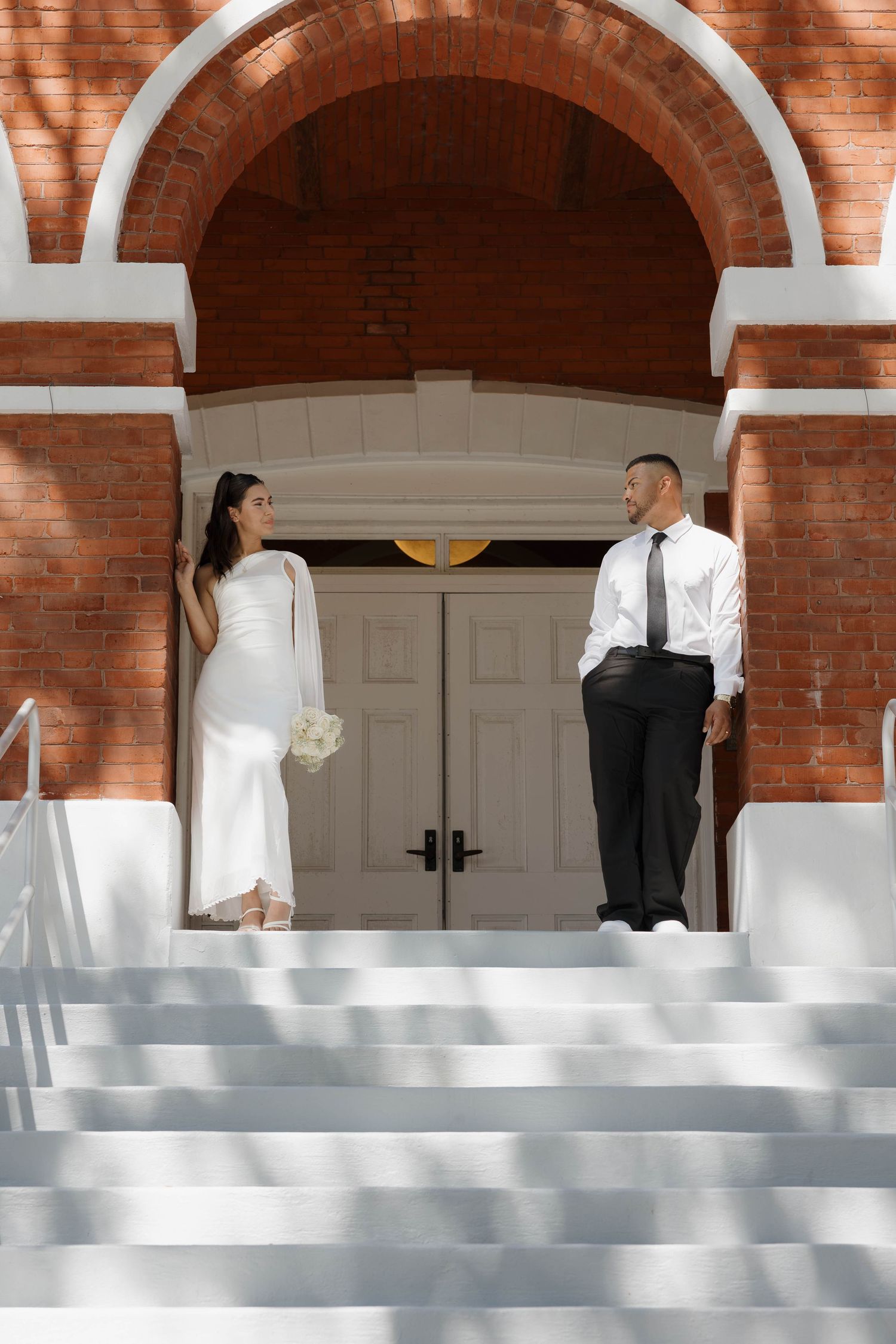 Bride and groom standing on white church steps under brick arch, bride holding white bouquet, sharing a glance.