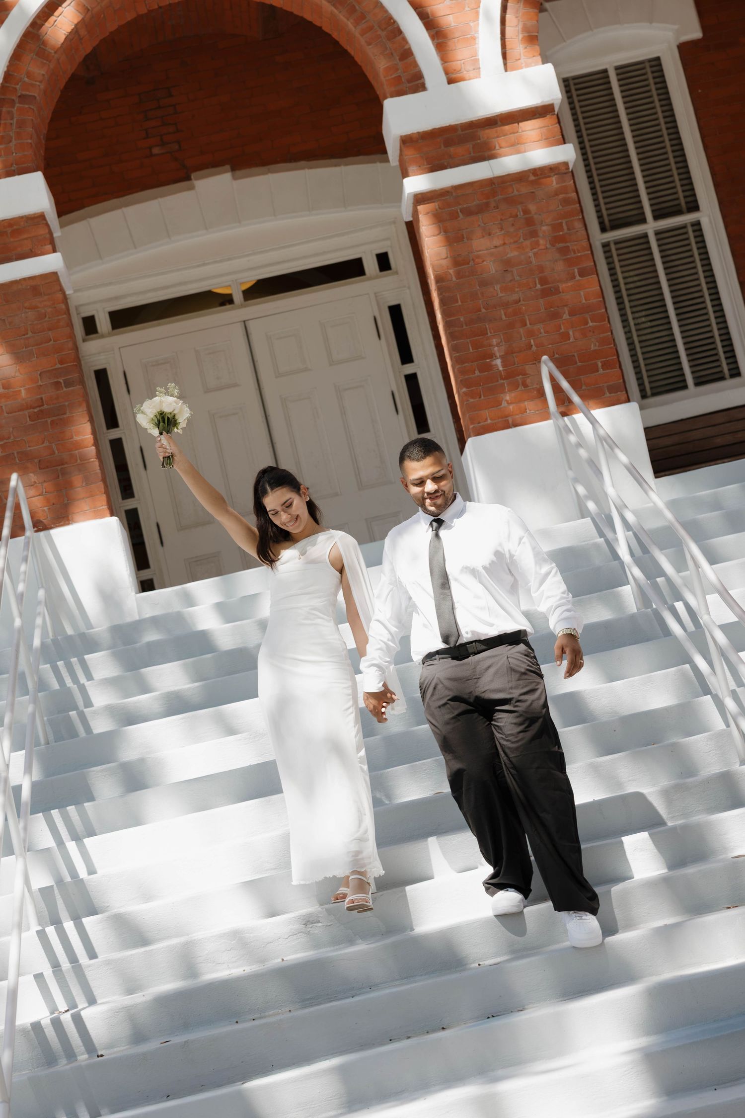 Newlywed couple descending white church steps, bride in white gown holding bouquet, groom in white shirt and tie.