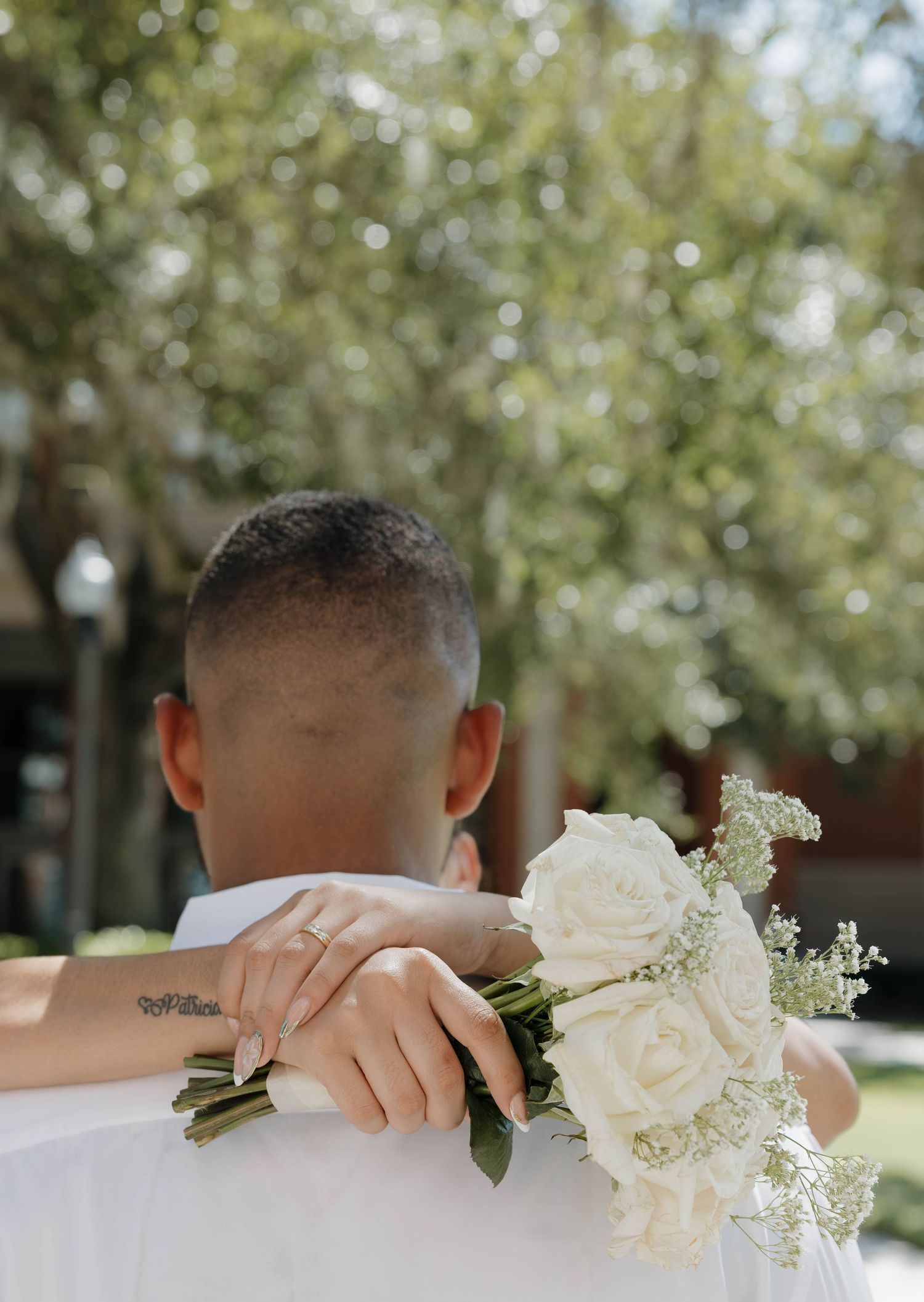 Bride rests her hands holding a white rose bouquet on the grooms shoulder from behind.