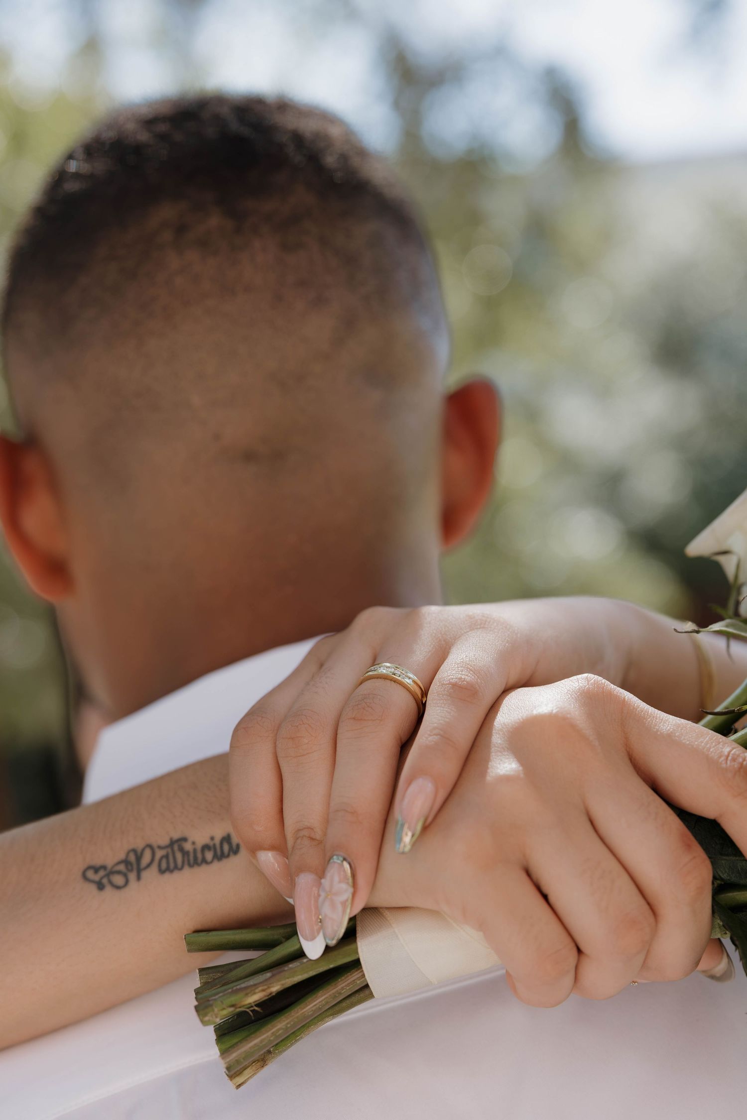 Close-up of bride and groom hands with gold wedding ring, bouquet, and Patricia name tattoo on wrist.