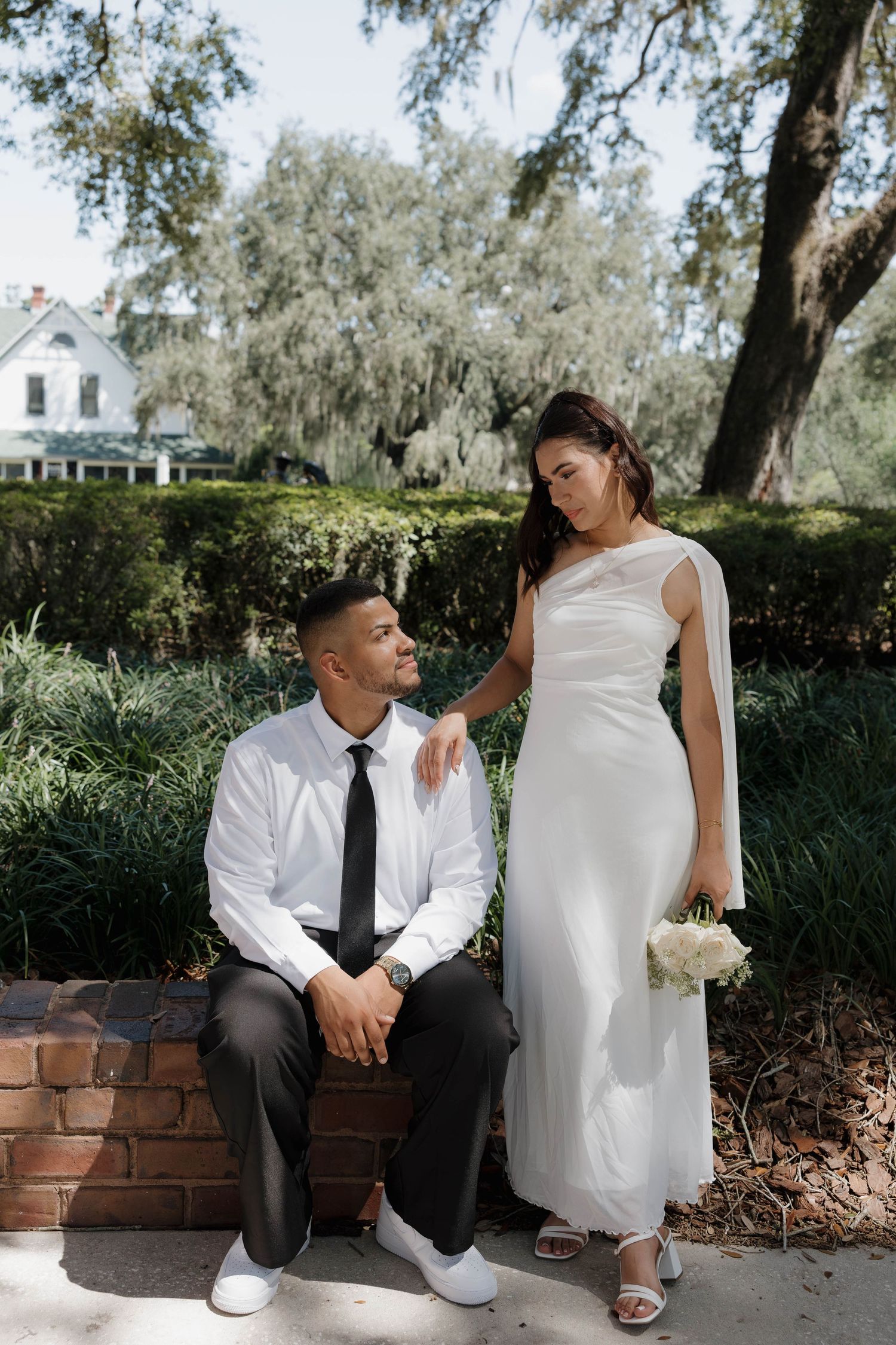 Bride in a white cape gown stands beside seated groom in a lush garden wedding portrait.
