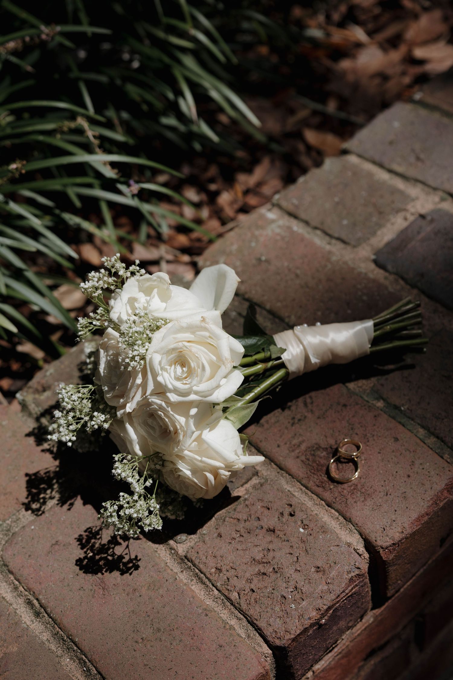 White rose bridal bouquet and two gold wedding rings rest on a rustic brick surface.