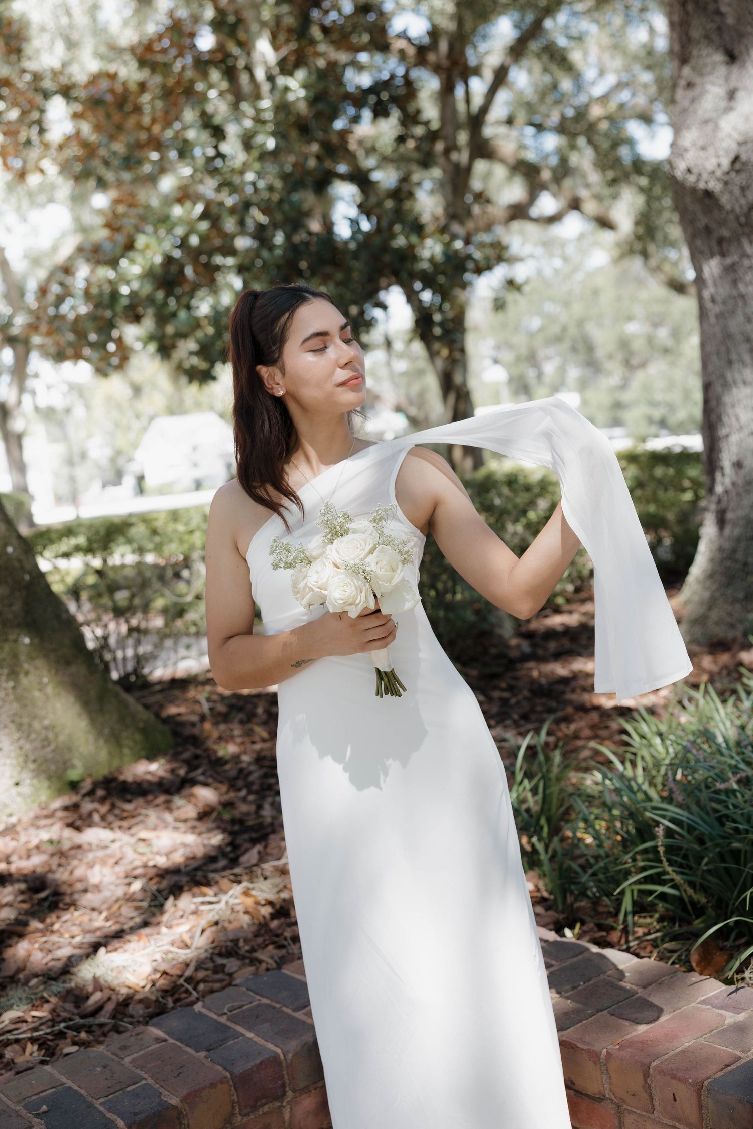 Bride in white gown holds bouquet and flowing ribbon outdoors among trees.