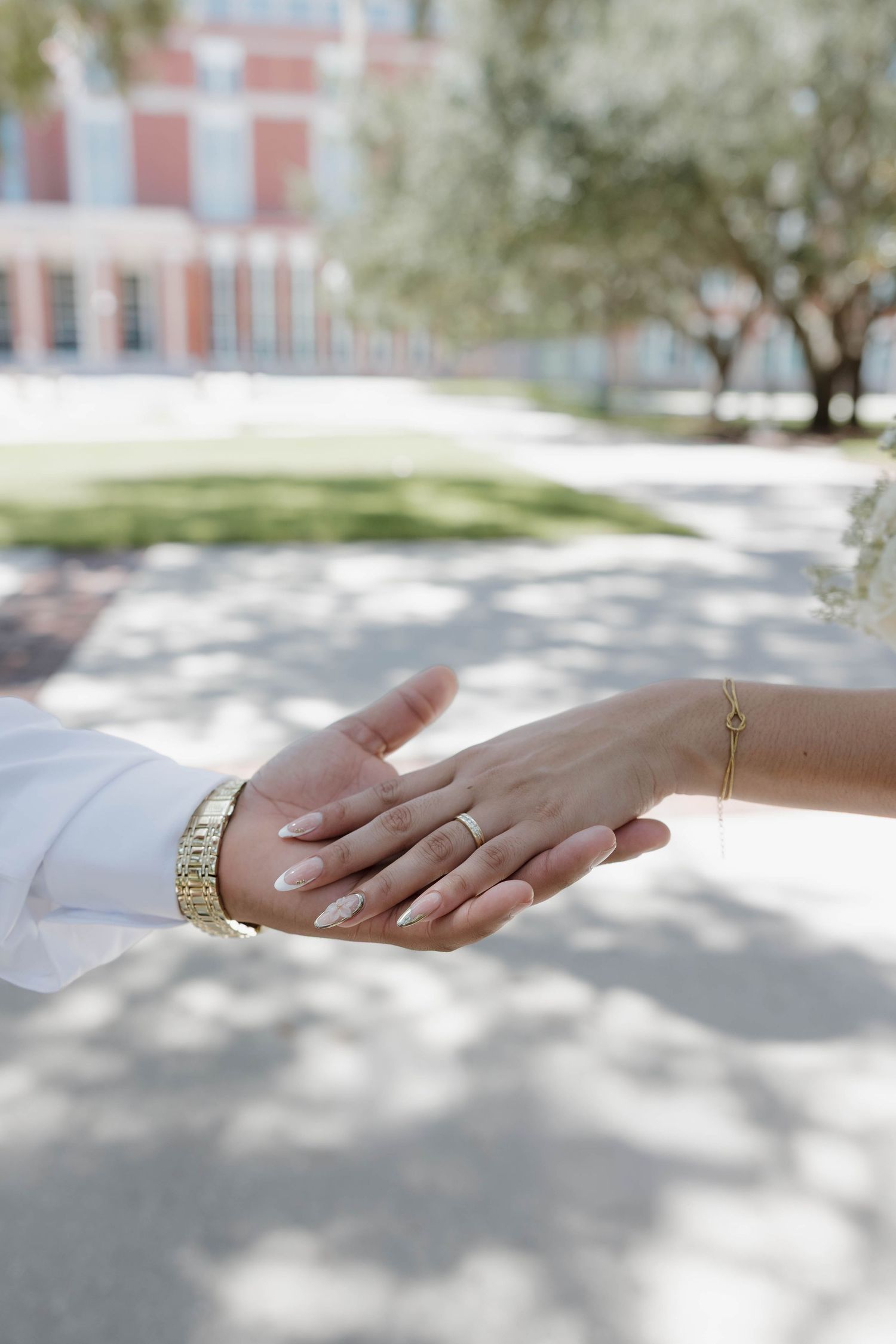 Close-up of bride and groom holding hands, showing gold bracelet and wedding ring on sunny outdoor pavement.