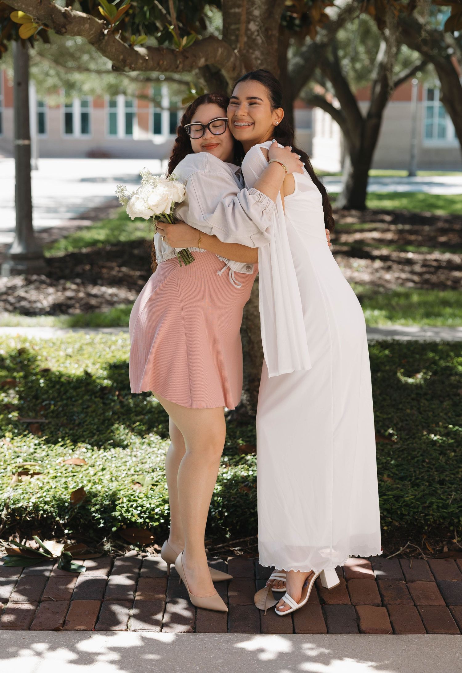 Bride in white dress hugging bridesmaid in pink skirt holding white bouquet outdoors.