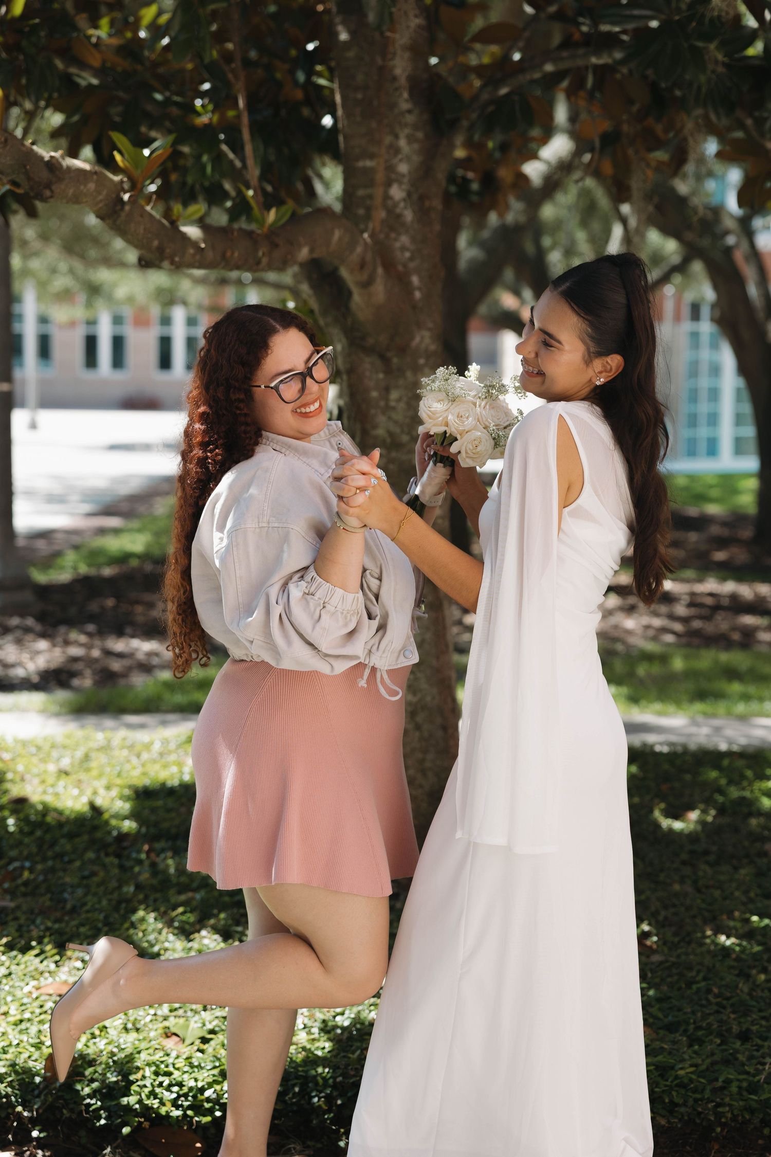 Joyful bride in white gown celebrates with a smiling bridesmaid holding a white bouquet outdoors.