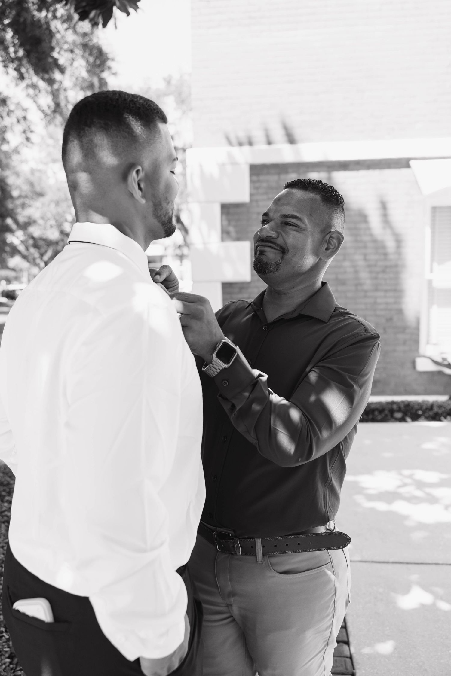 Black and white photo of two men outdoors, one adjusting the other's tie or collar before a formal event.