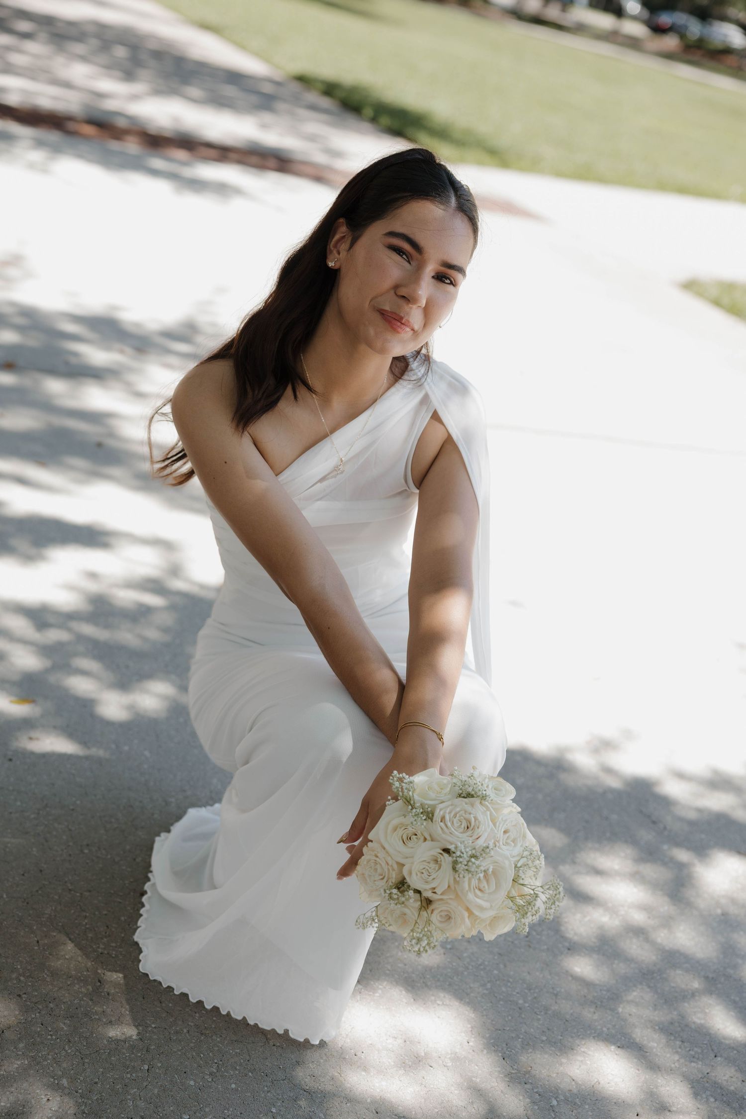 Bride in elegant white one-shoulder gown poses outdoors holding bouquet of white roses on sunny day.
