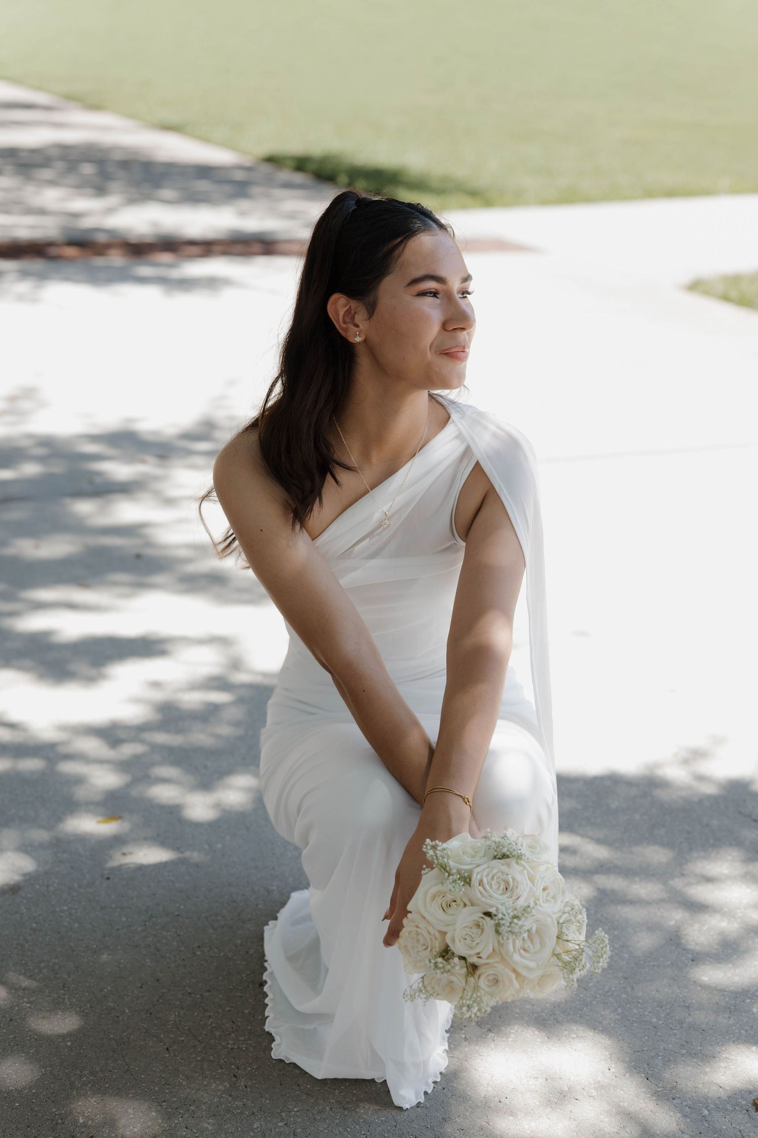 Bride in white one-shoulder gown kneeling outdoors, holding bouquet of white roses, smiling softly in natural light.
