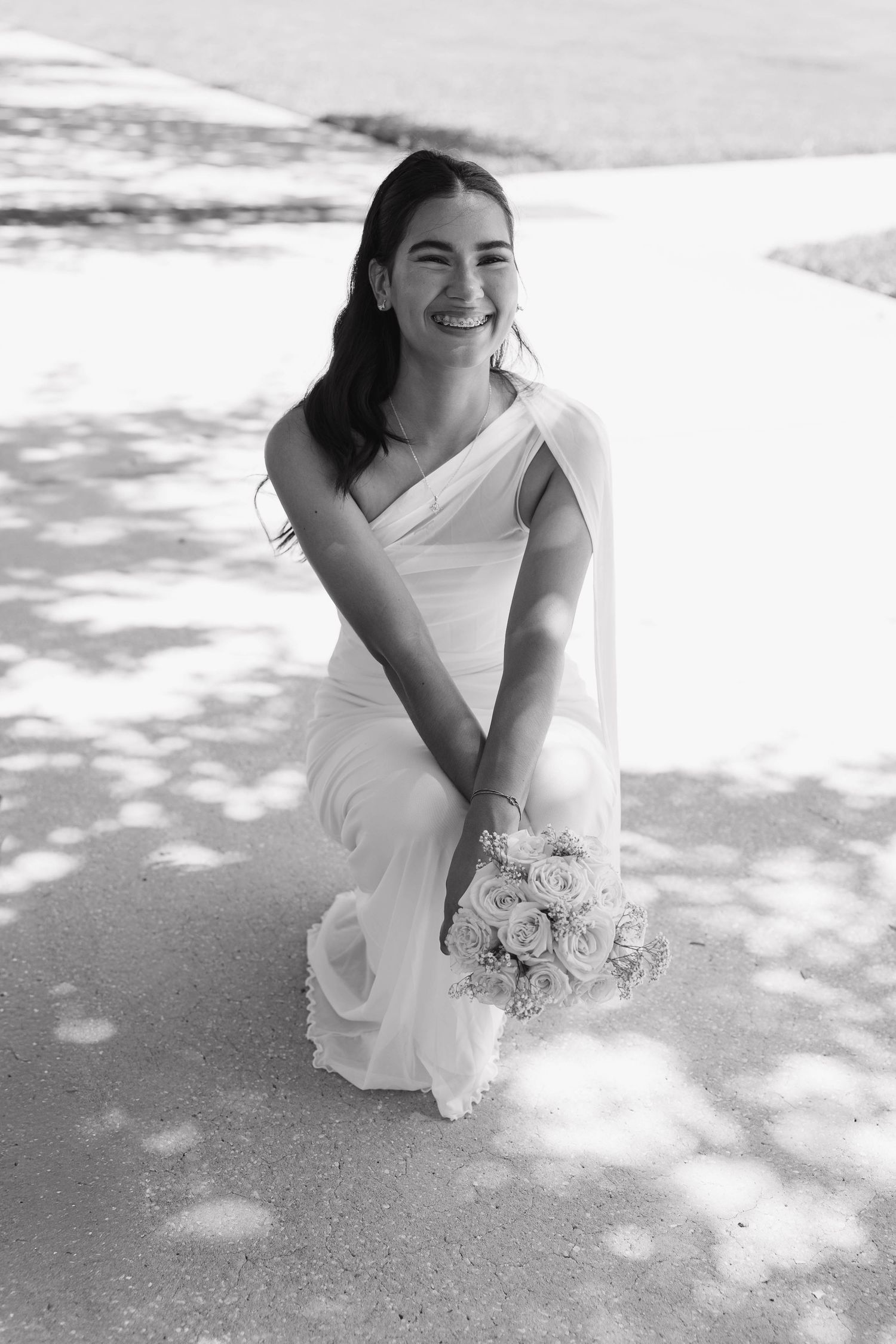 Smiling bride in white one-shoulder gown kneels outdoors holding rose bouquet, black and white photo.