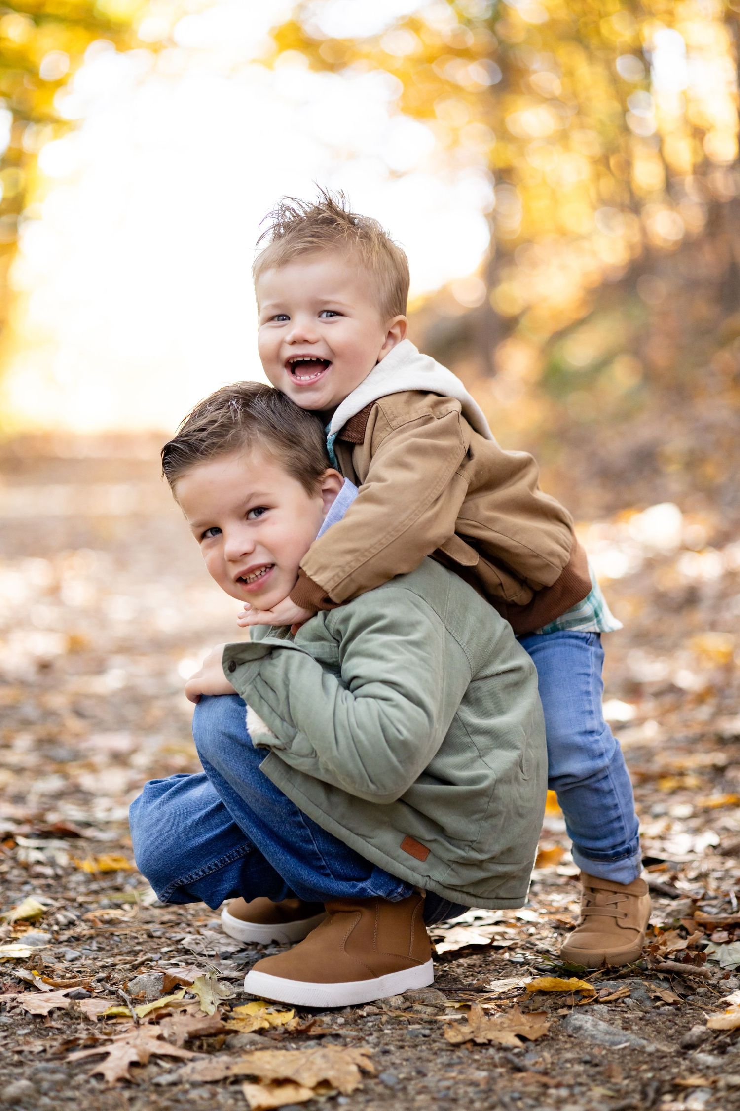 Two young boys in autumn jackets smile and play together on a leaf-covered path surrounded by fall foliage.