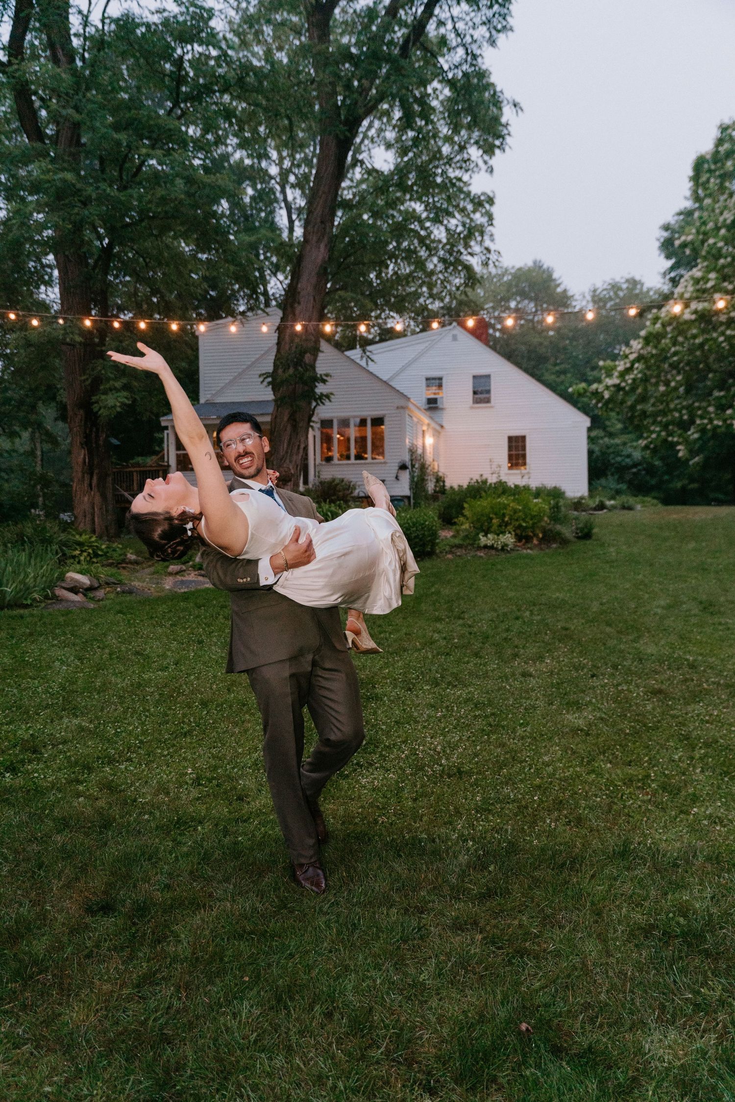 Groom lifts bride in backyard wedding celebration at dusk with string lights and white farmhouse backdrop.
