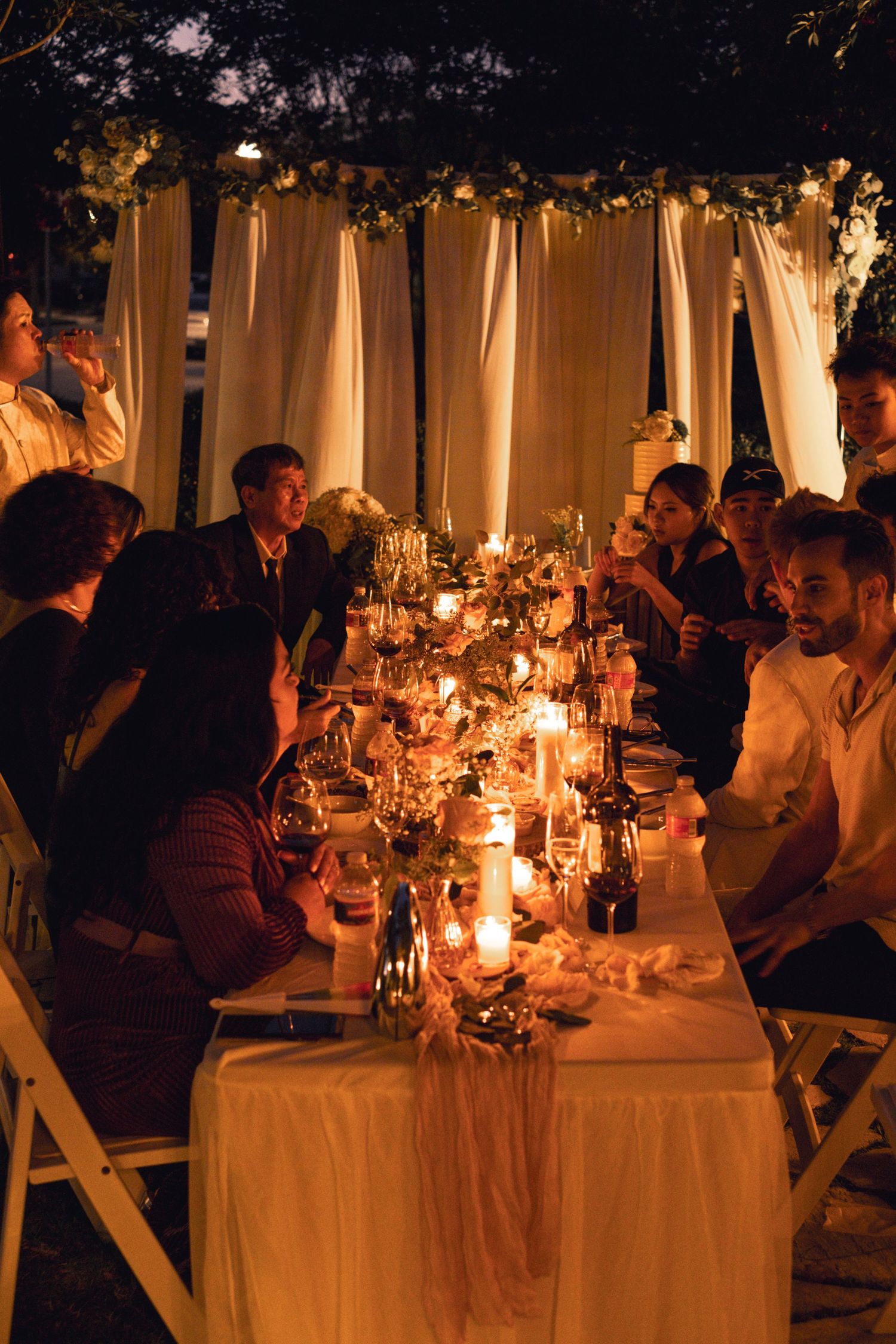 Guests gathered around candlelit outdoor wedding reception dinner table at night.
