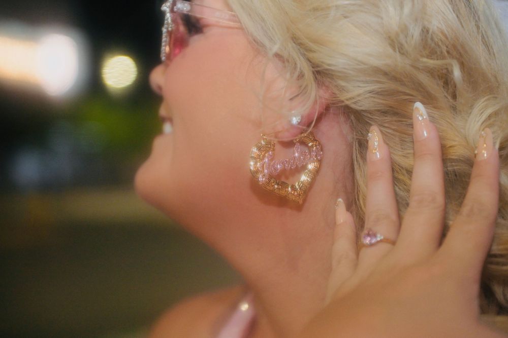 Close-up of a woman wearing a large pink heart-shaped glitter earring with manicured nails and pink sunglasses.
