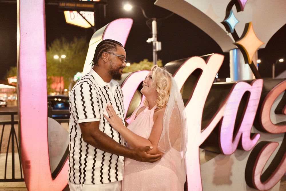 Newlywed couple posing in front of glowing Las Vegas sign at night, bride in veil and groom in patterned shirt.