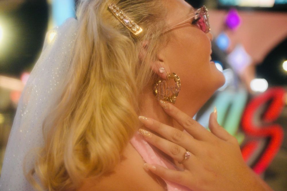 Blonde woman wearing pink sunglasses, veil, hoop earrings, and pink outfit at a vibrant nightlife venue.