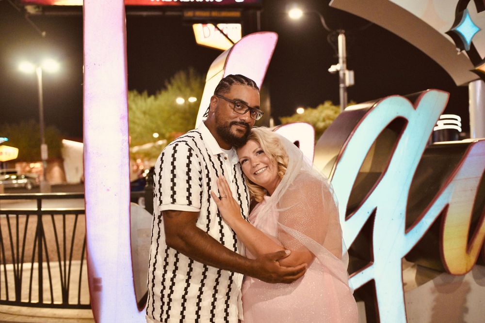 Couple embracing and smiling outdoors at night near illuminated signs at a Las Vegas venue.