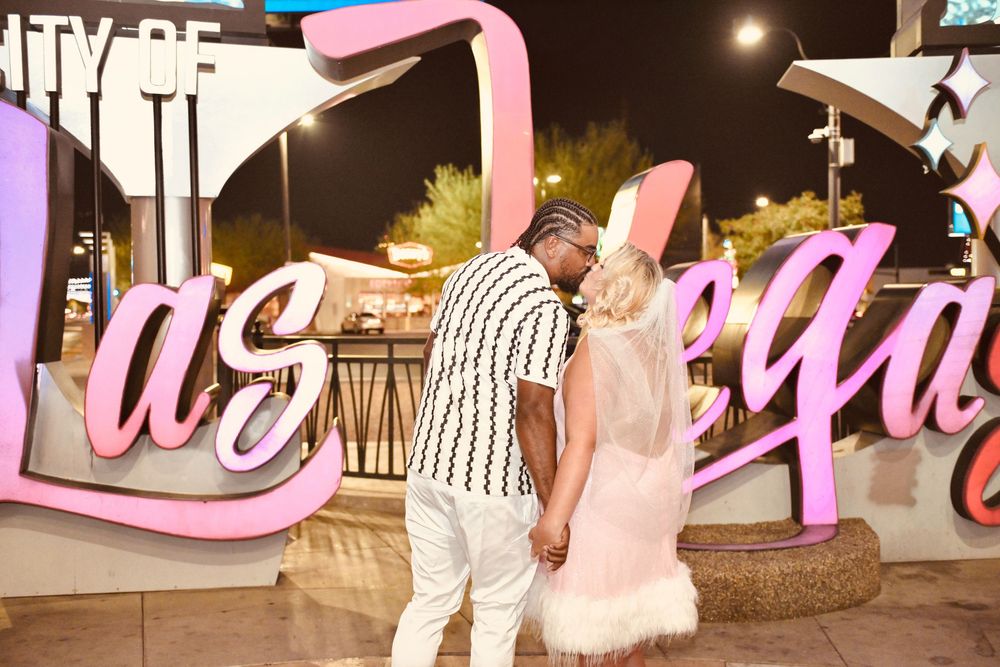 Couple holding hands in front of the Las Vegas sign at night, woman in pink bunny costume, man in striped outfit.