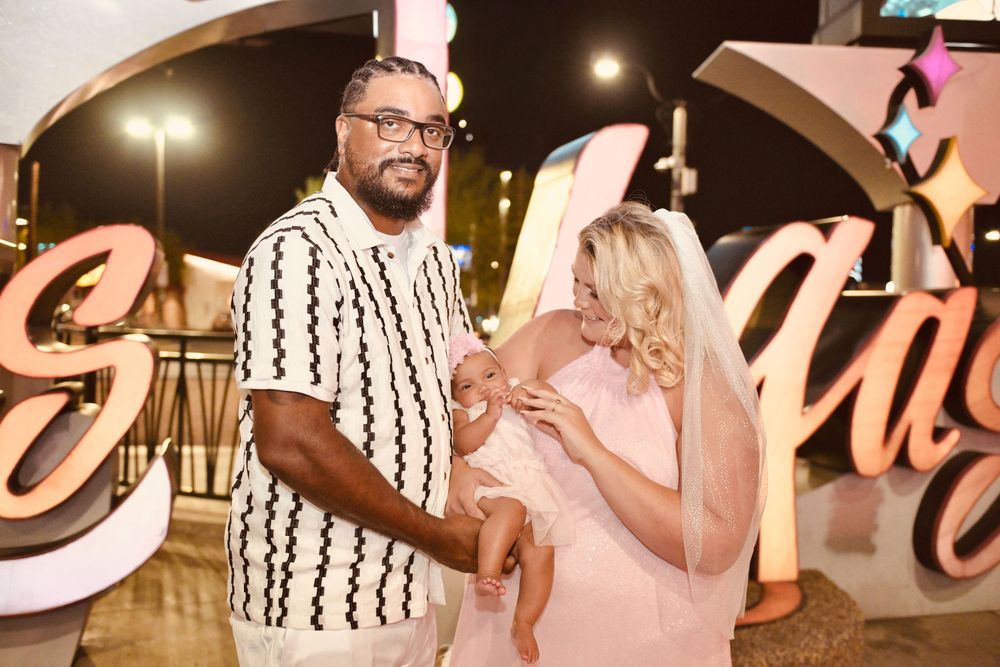 A couple with a baby poses in front of decorative Las Vegas signs at night.