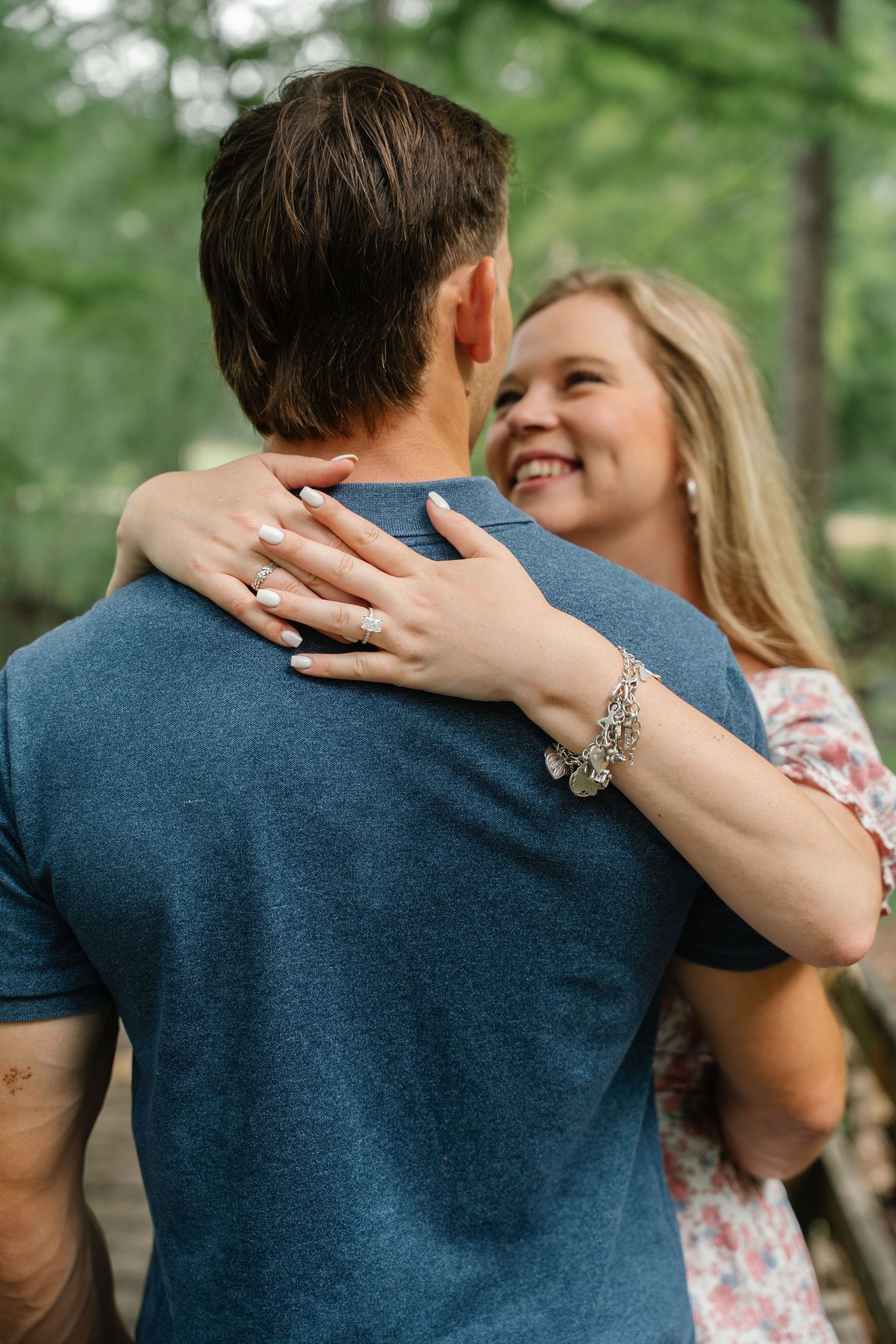 Smiling blonde woman hugging a man in a blue t-shirt outdoors, showcasing an engagement ring and charm bracelet.