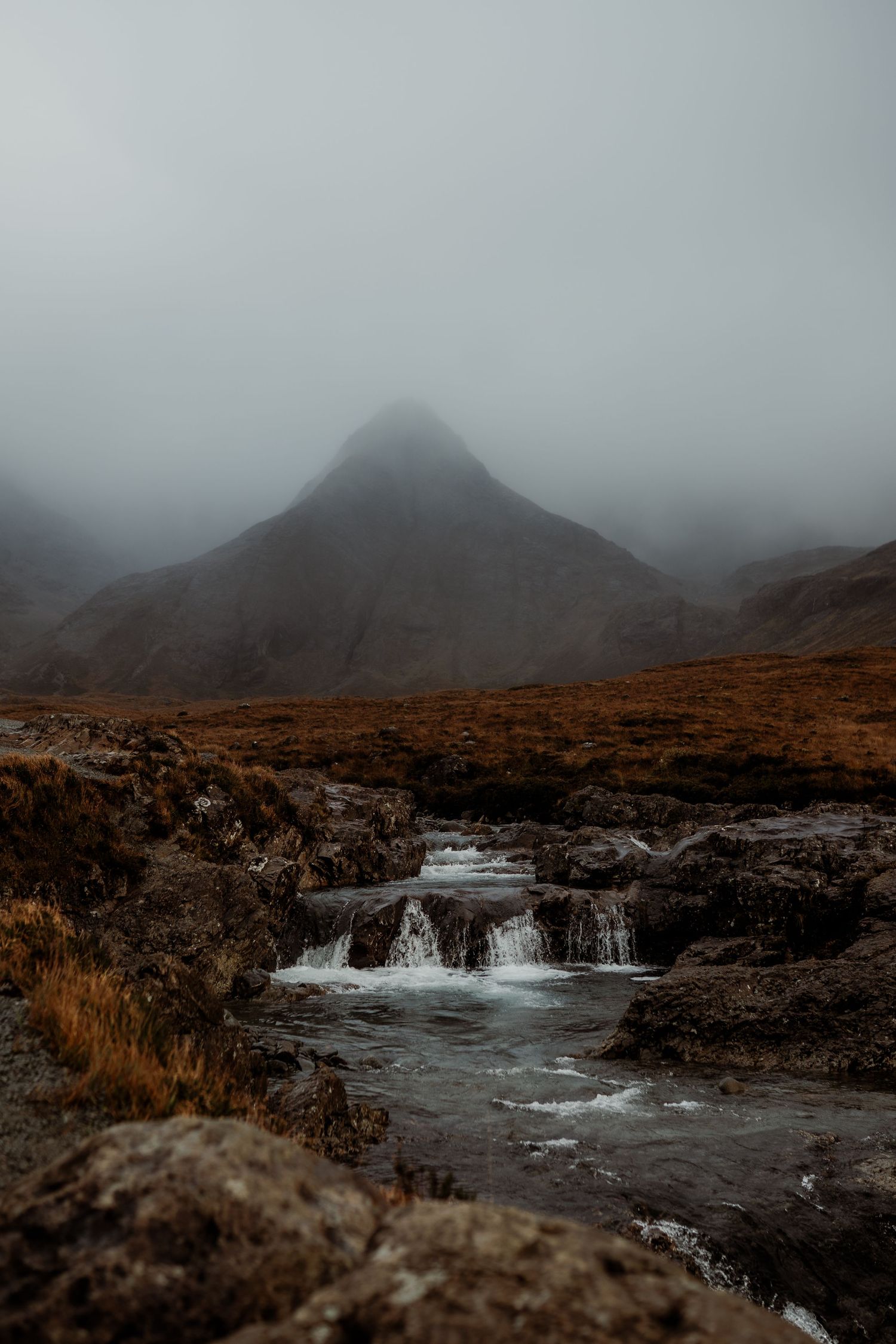 Misty mountain landscape with a small waterfall cascading over rocks into a stream on a foggy, overcast day.