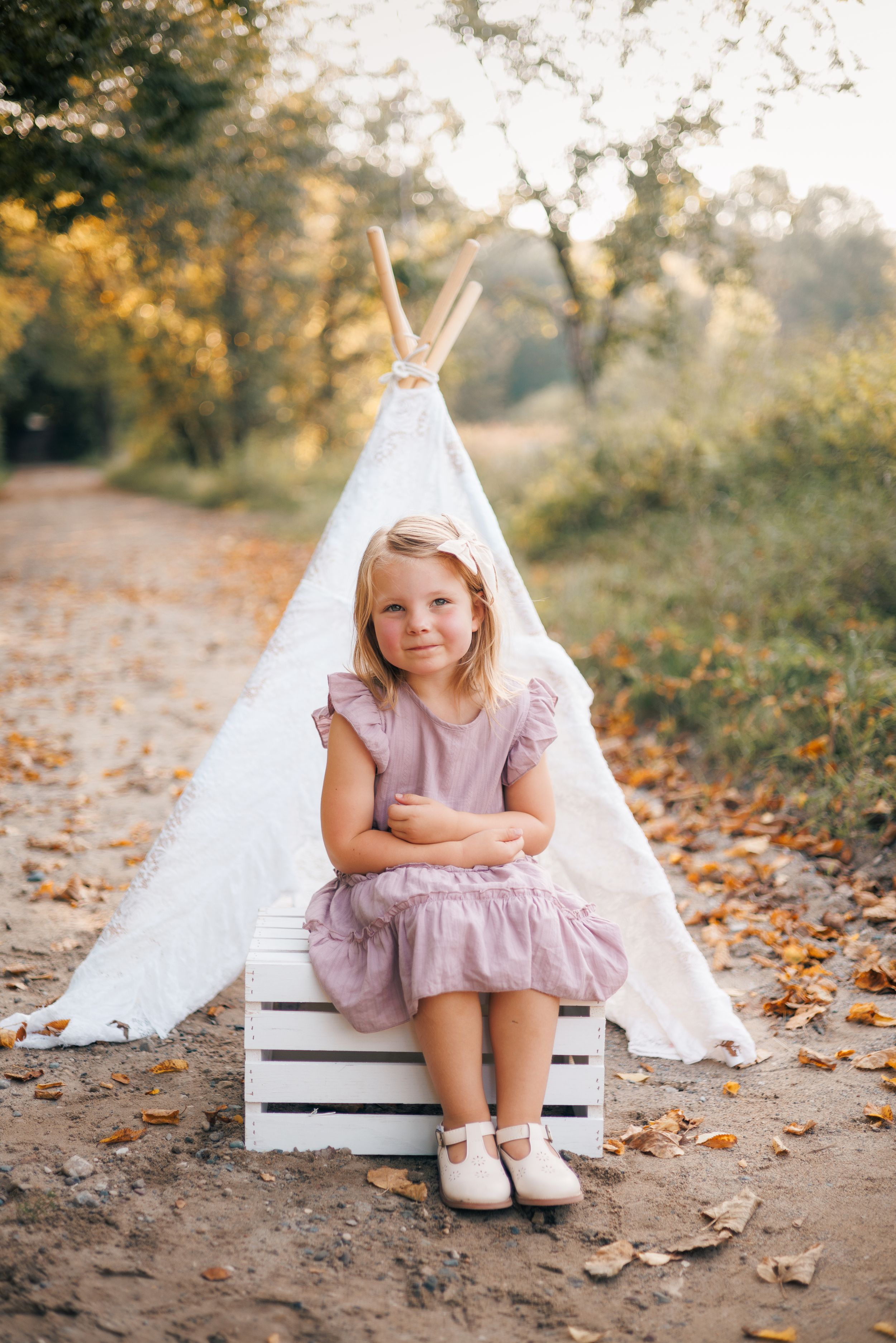Young blonde girl in pink dress sitting on white wooden crate before teepee tent in autumn forest setting.