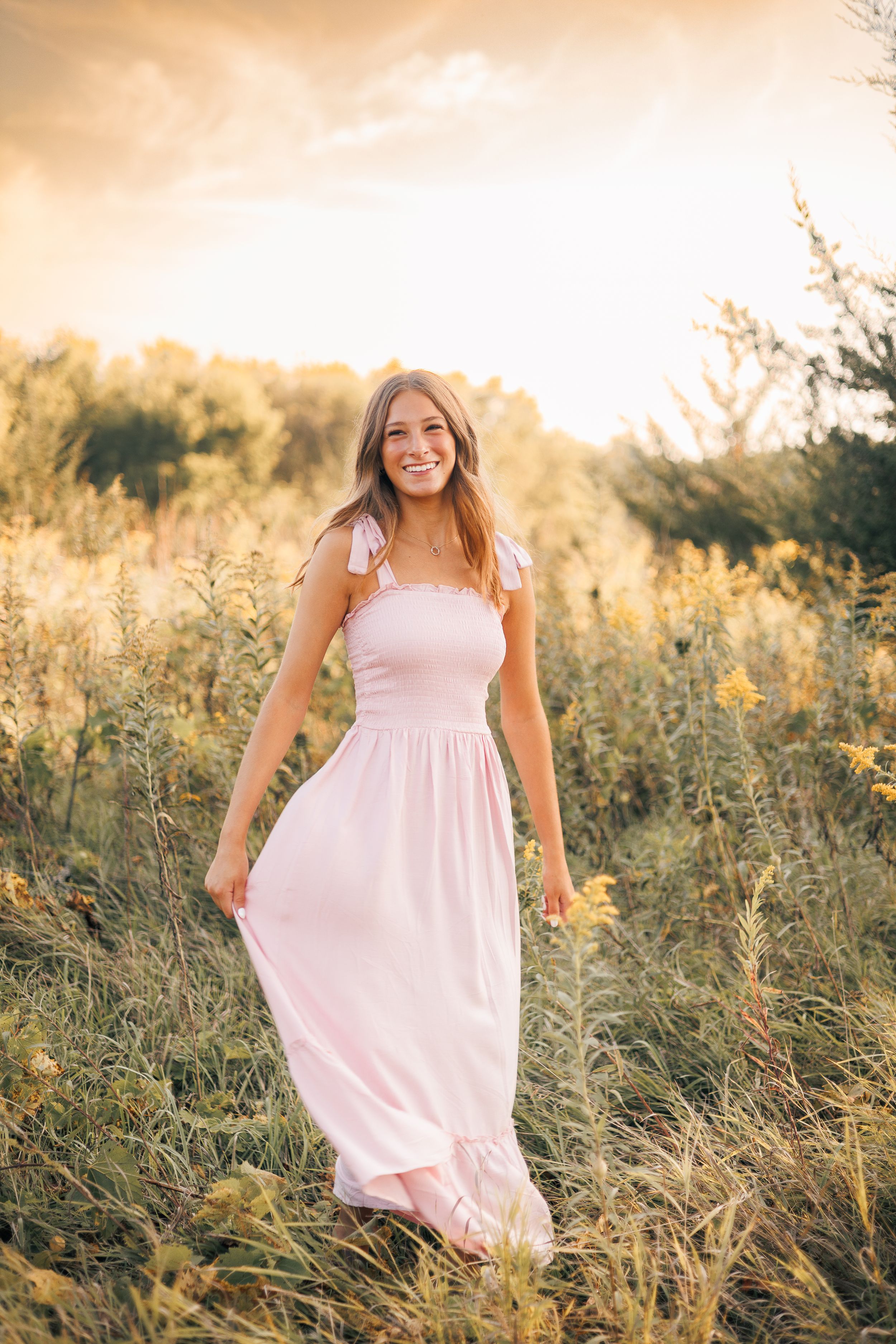 Young woman in a pink maxi dress smiling in a golden sunlit field of wildflowers.
