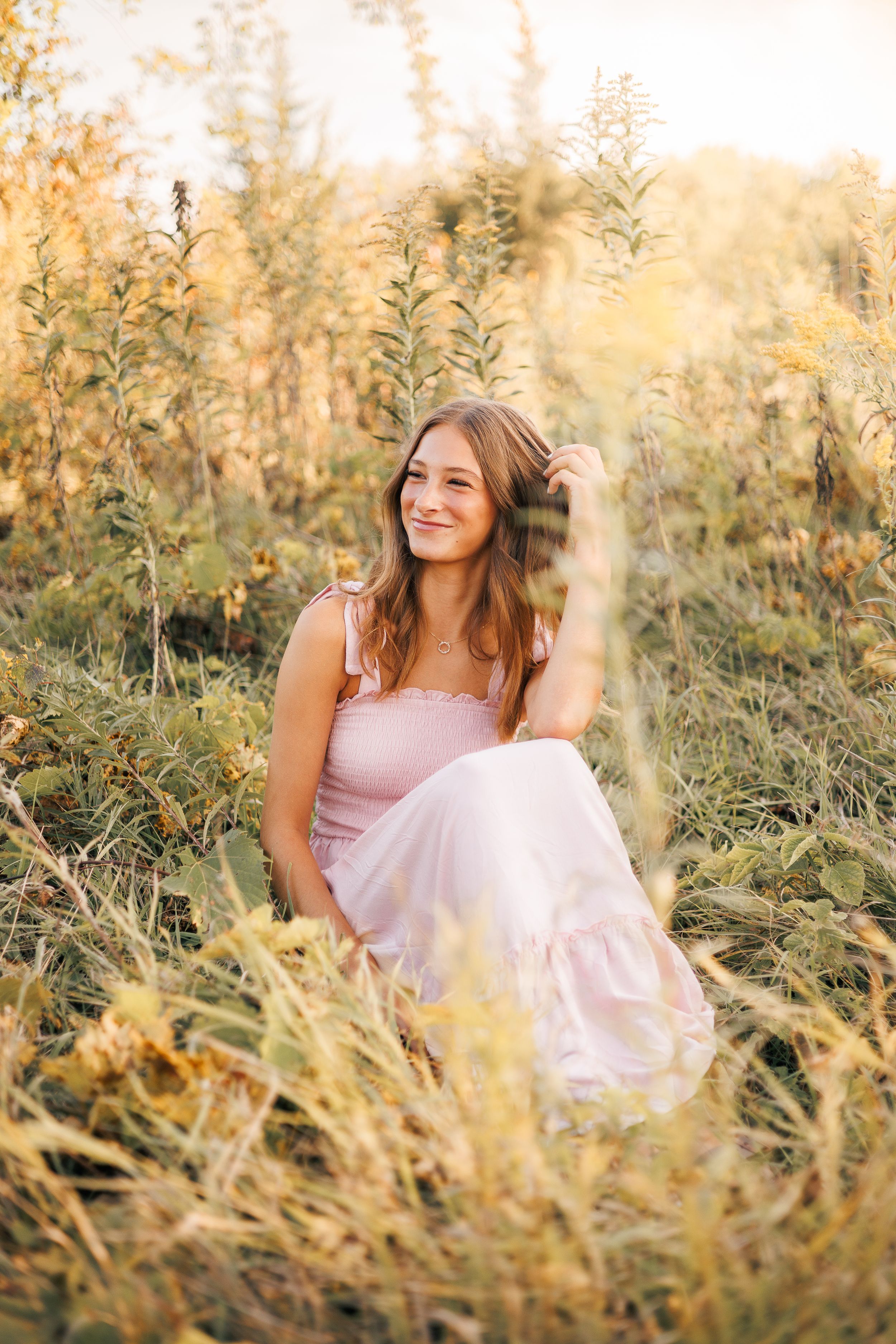 Young woman in pink dress sitting among golden wildflowers in a sunlit field, smiling warmly.