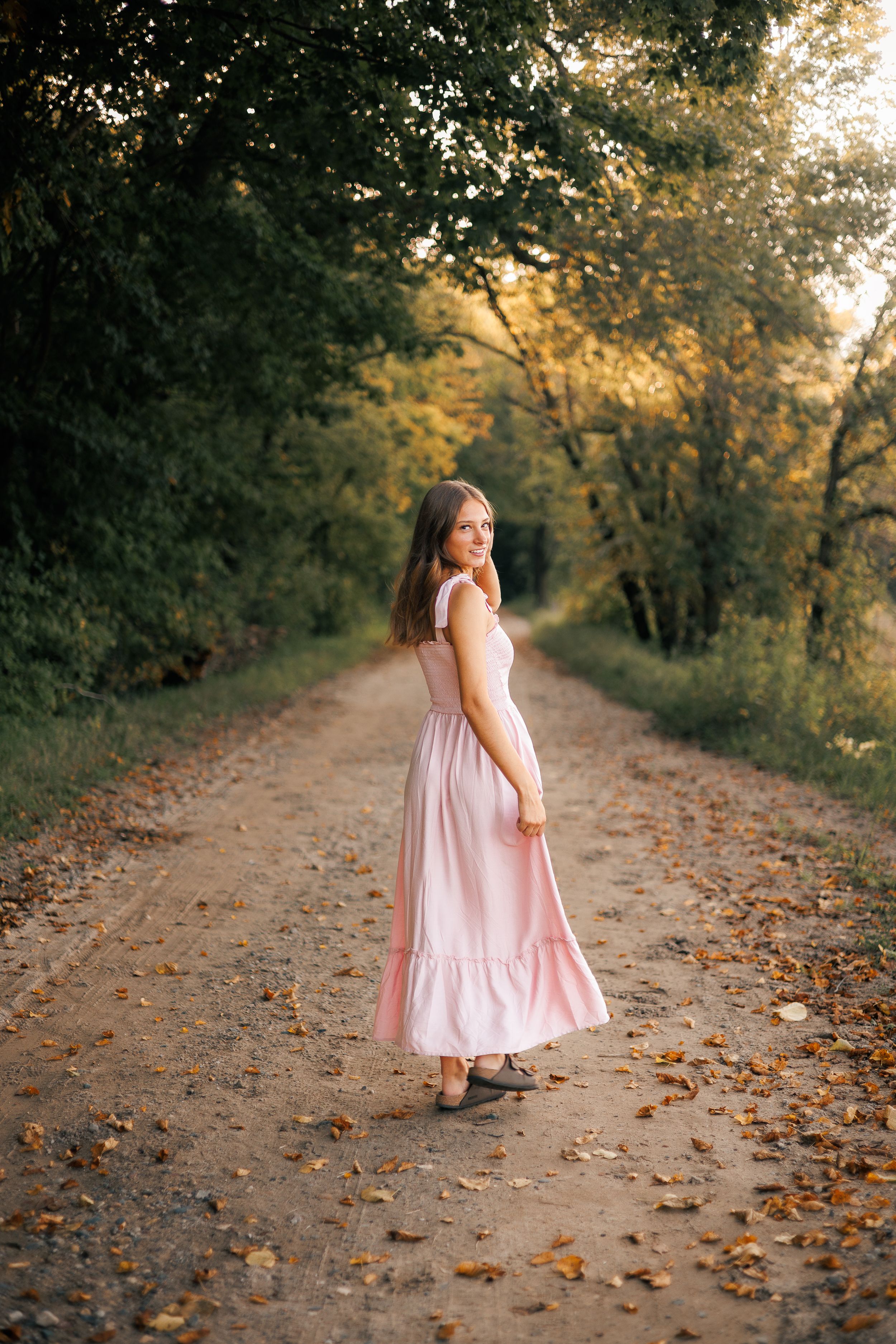 Young woman in pink maxi dress walking on a tree-lined dirt path during golden hour sunset.
