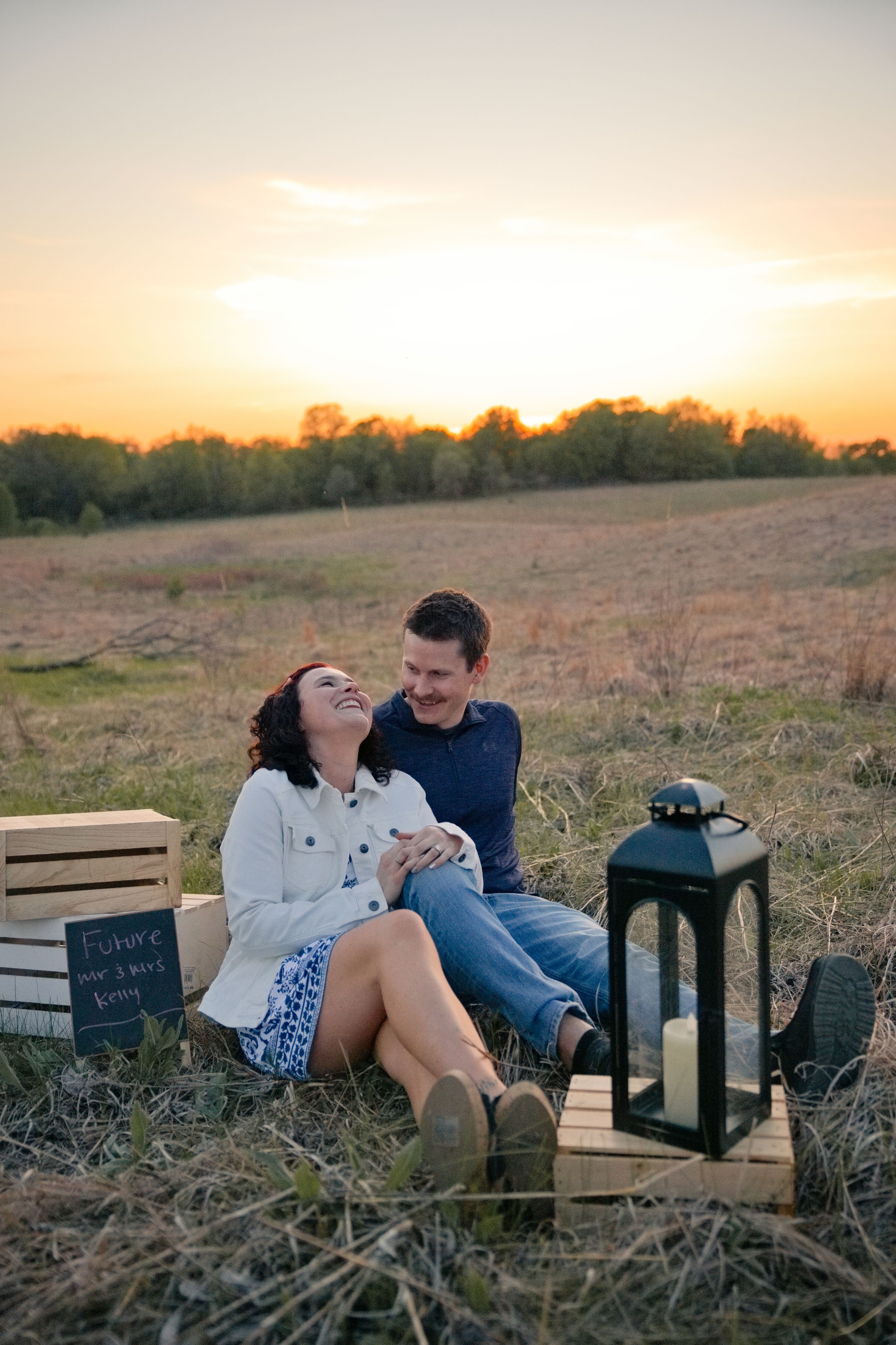 Couple sitting together in a golden field at sunset, with a lantern and wooden crates nearby.