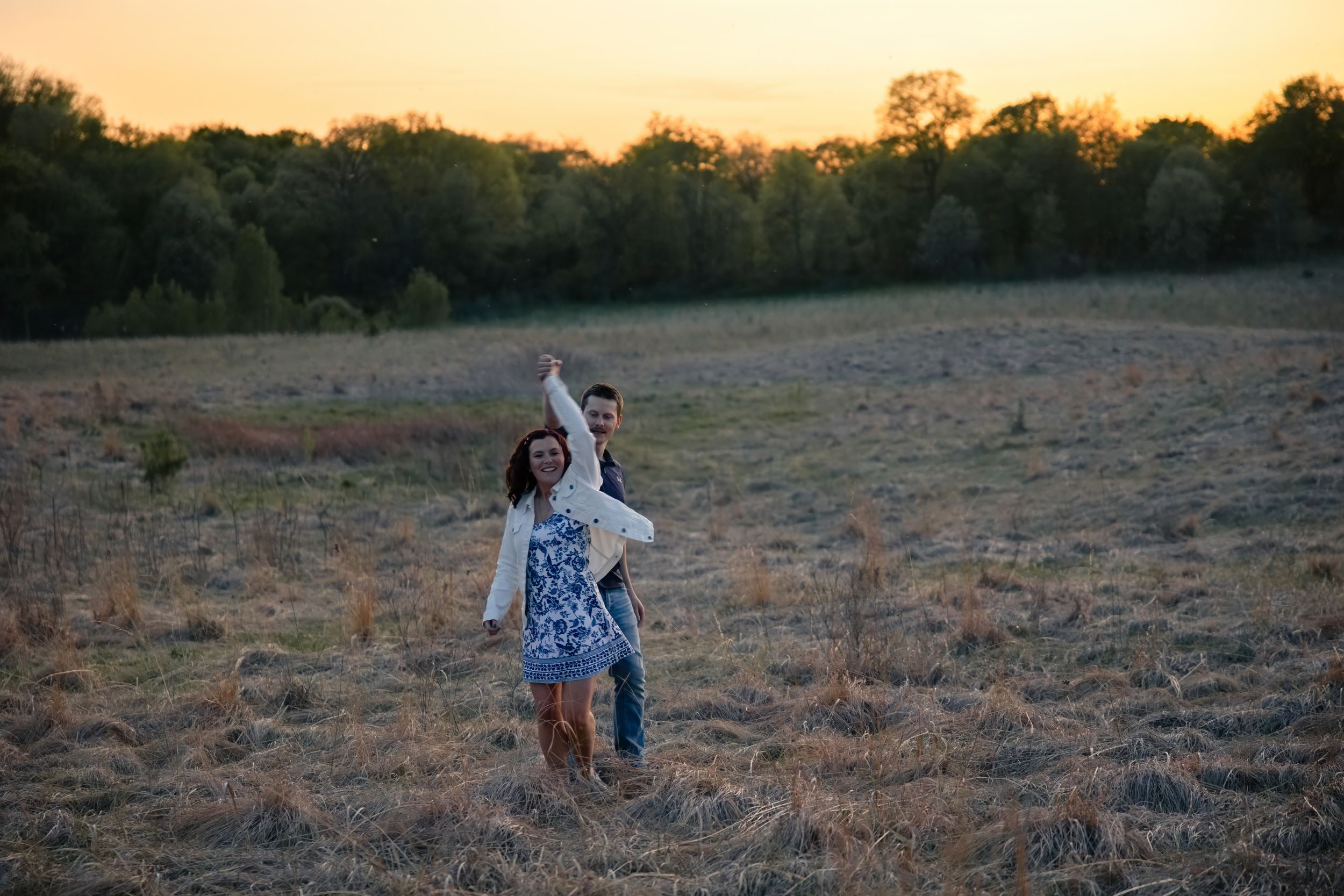 Couple dancing joyfully in a golden field at sunset with trees in the background.