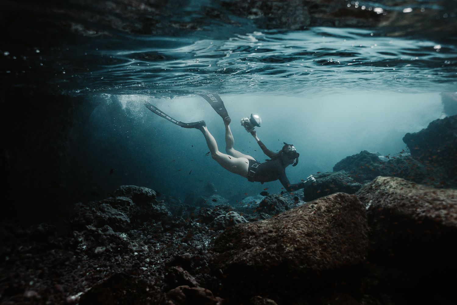 Freediver exploring underwater rocky cave with dramatic light rays filtering through the surface.