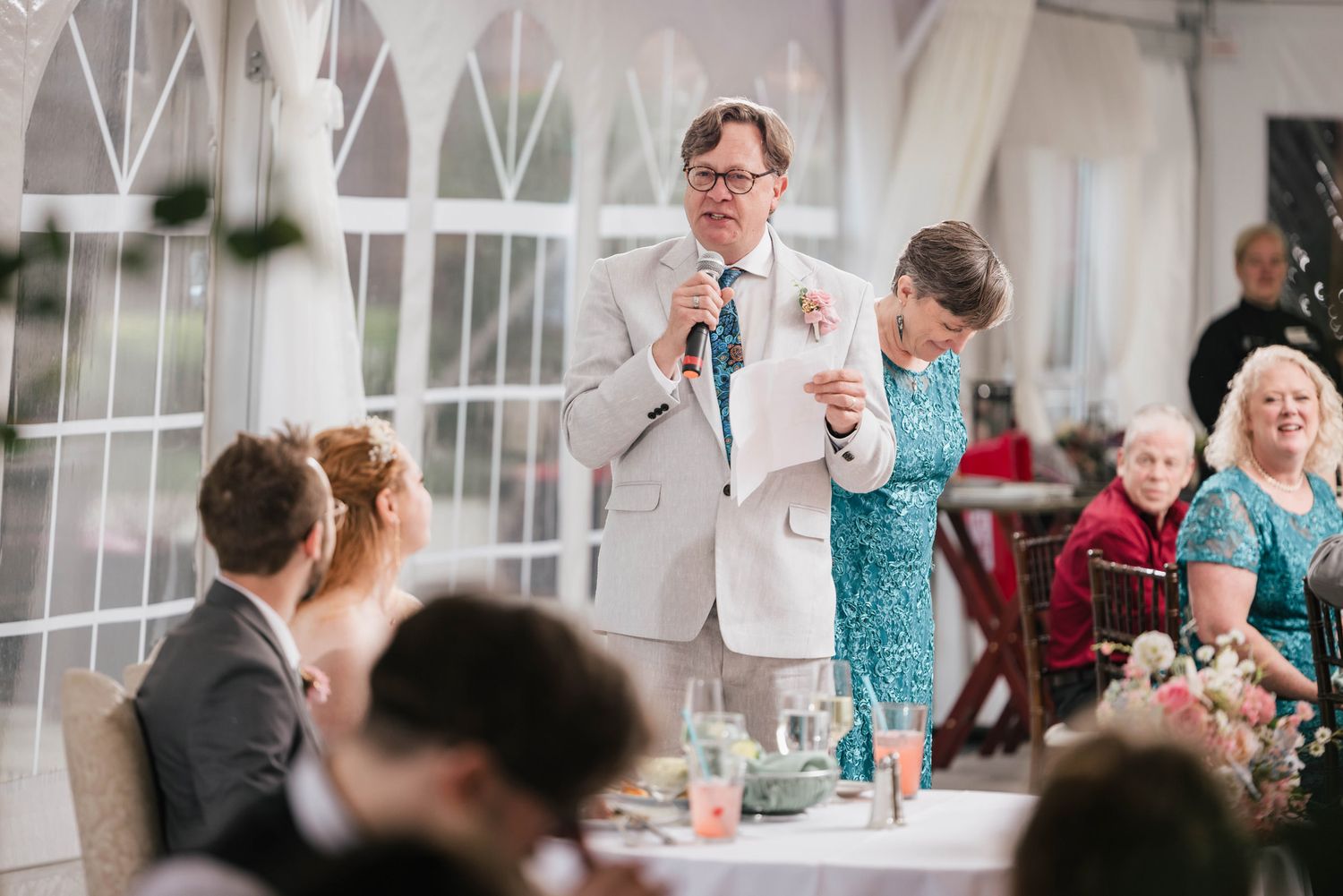 Groom's father giving a speech, holding his notes, groom's mother standing beside him.