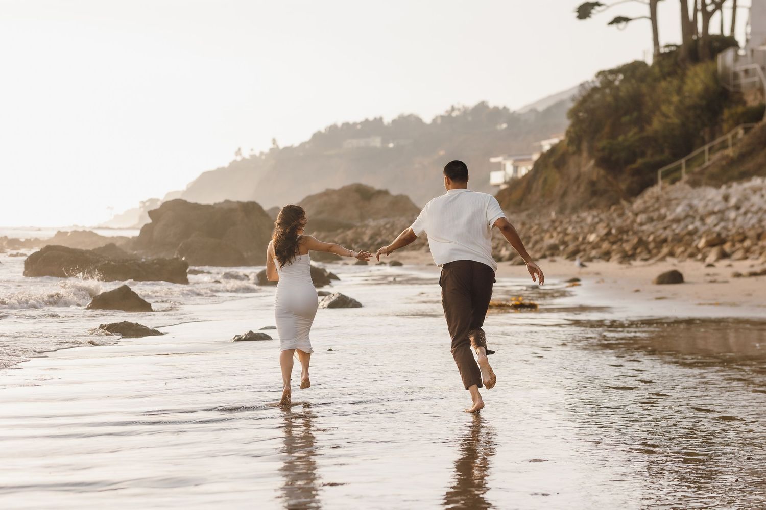 Couple running barefoot along a sunlit beach with rocky cliffs, holding hands in a romantic coastal setting.