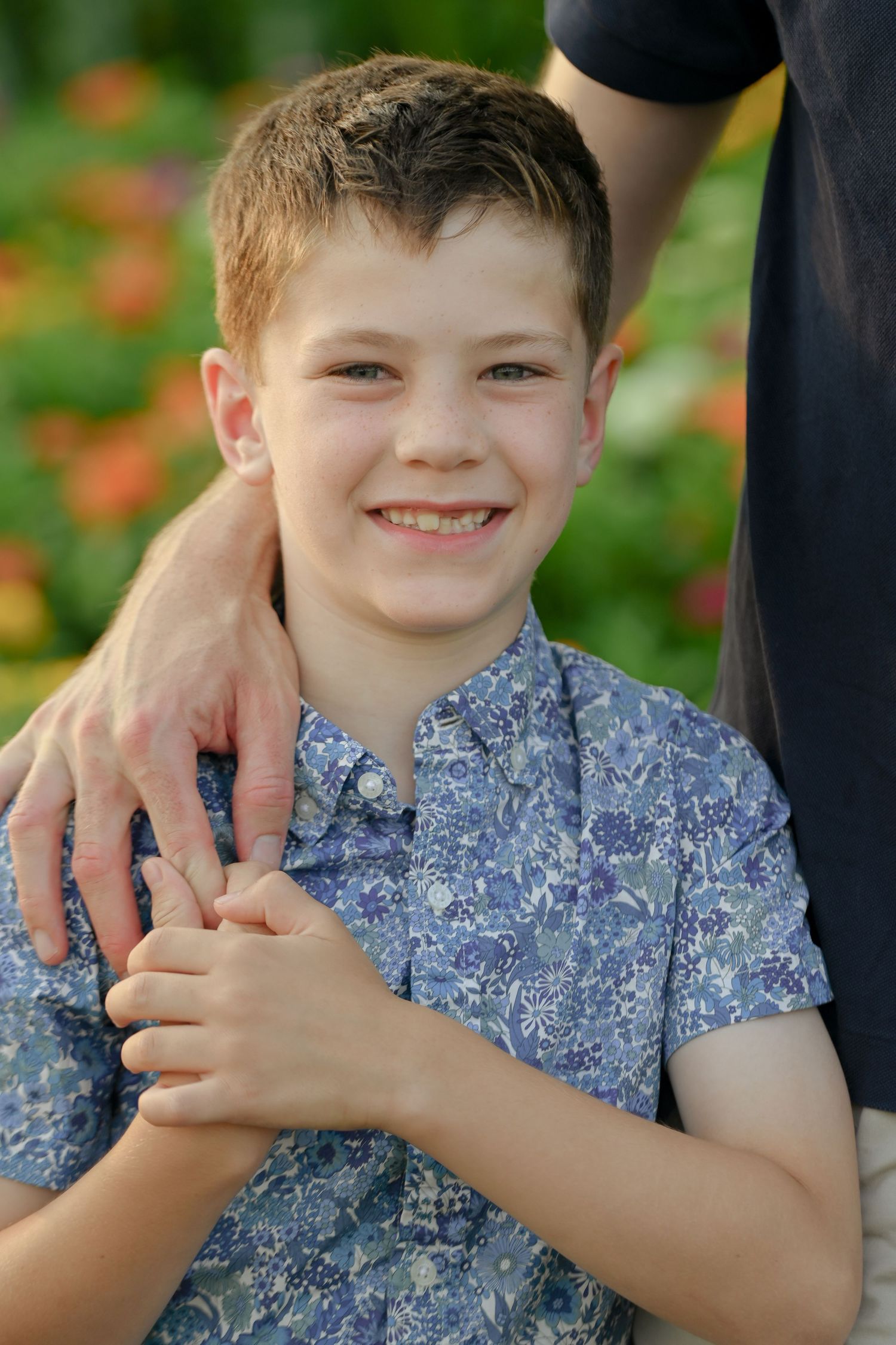 Smiling boy in floral blue shirt holding an adult's hand outdoors with colorful flowers in background.