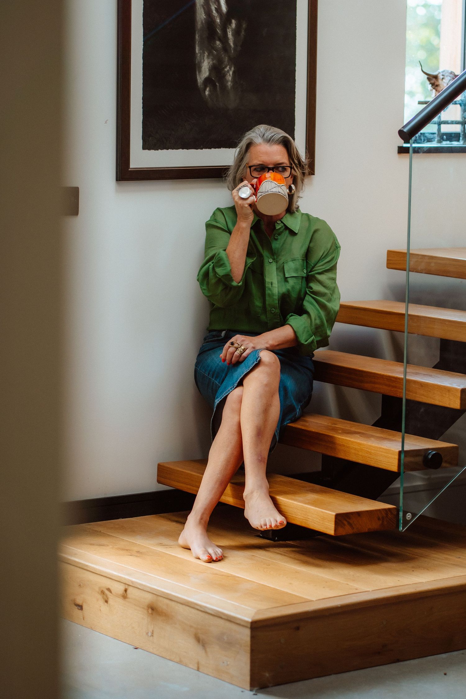 Woman in green shirt sitting barefoot on wooden stairs, drinking from a colorful mug in a modern home.